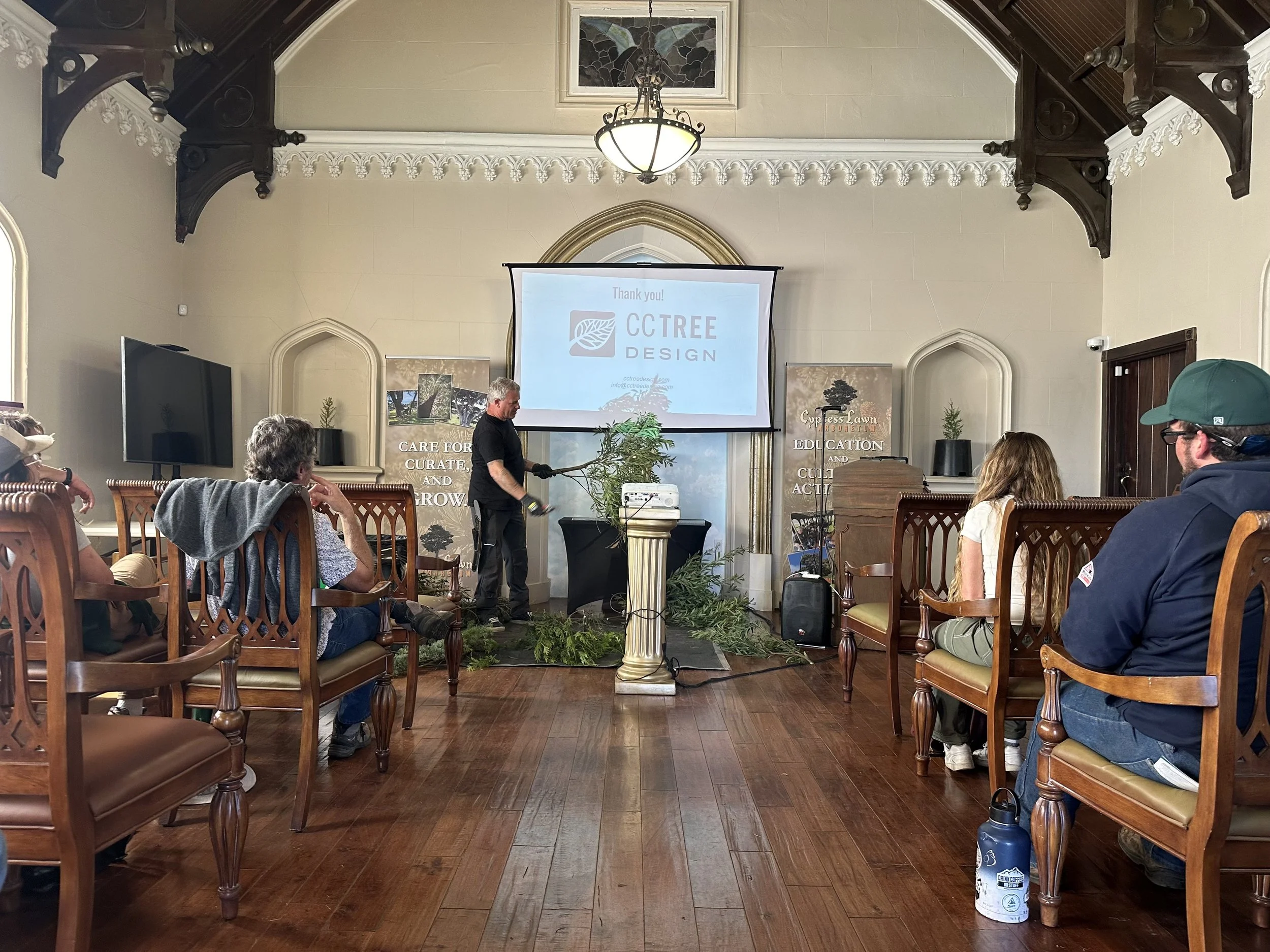 A presentation in a room with a wooden floor, several attendees sitting on wooden chairs, and a man standing near the front holding pruning shears and branches. A large screen displays the logo 'CCTREE Design,' and there are banners on either side of