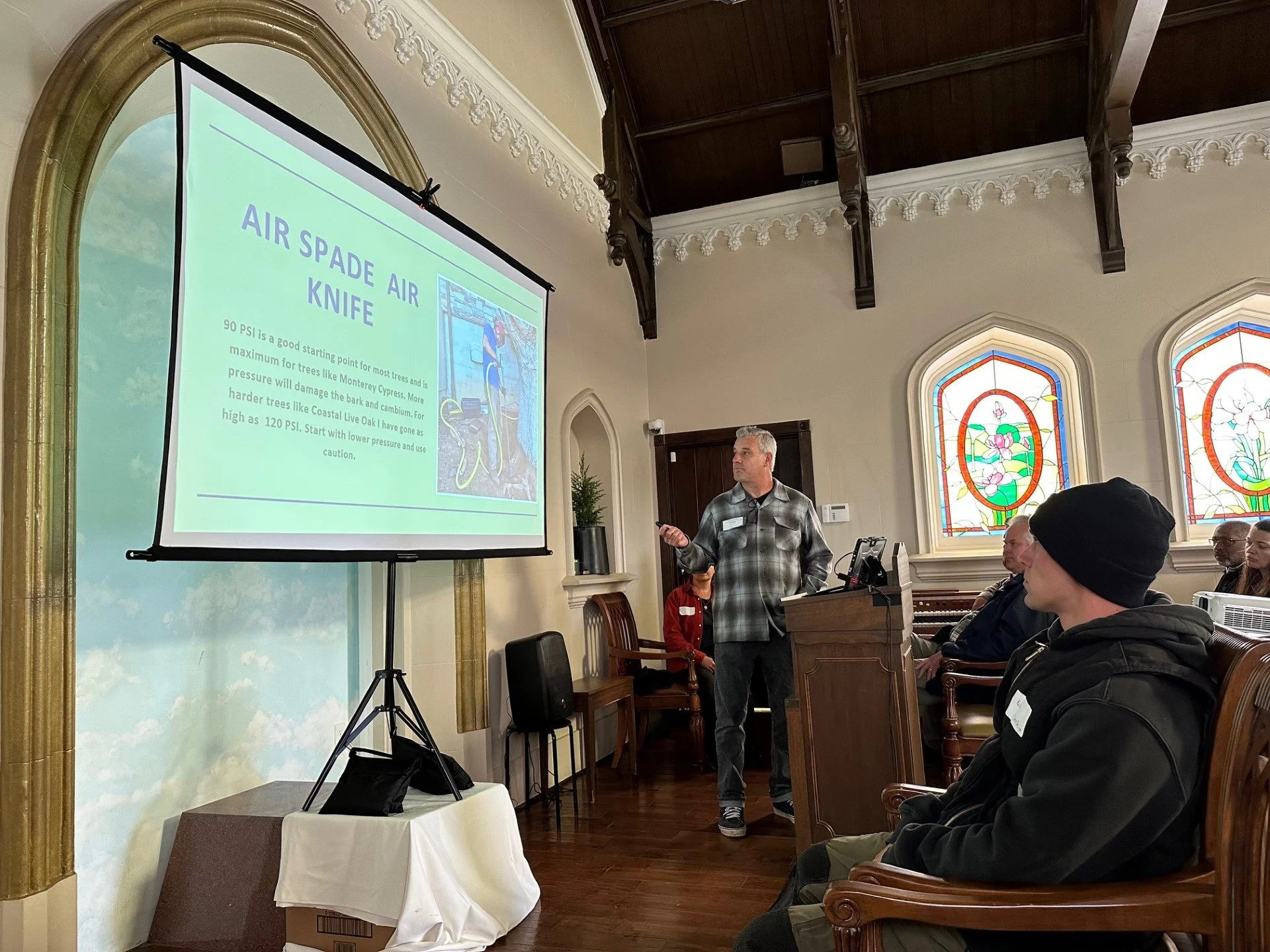 A man giving a presentation on an air spade air knife in a room with stained glass windows. Several audience members are seated, listening to the speaker.