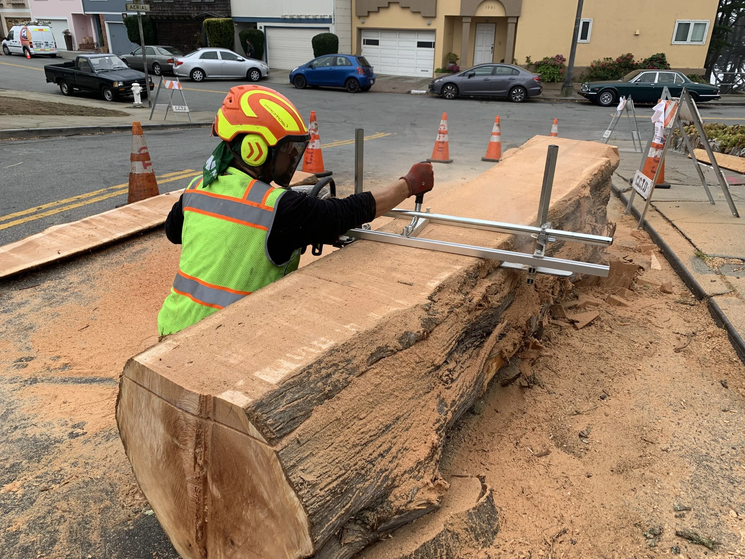 A construction worker with a yellow and red safety helmet, green vest, and gloves is cutting a large fallen tree trunk with a power saw on a city street. Orange cones and barricades surround the work area.