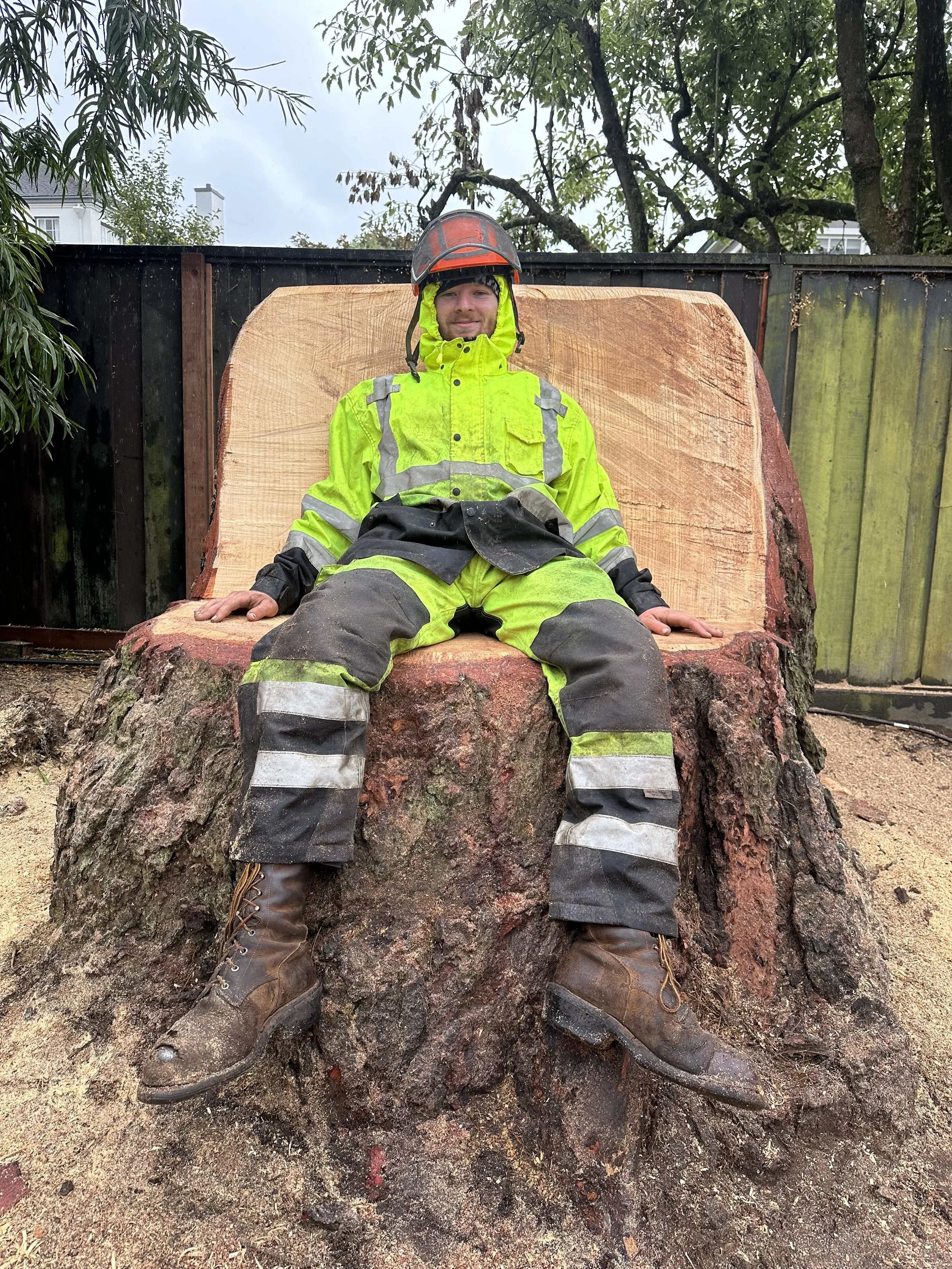 A man sitting on a large tree stump cut in half, wearing a bright yellow safety jacket, black and yellow protective pants, brown work boots, and a safety helmet, outdoors with trees and a wooden fence in the background.