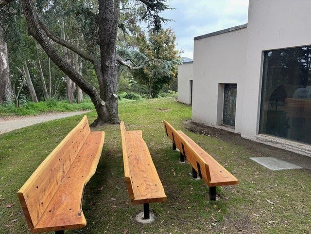 Two wooden park benches and a smaller wooden seat in front of a modern white building with large windows, surrounded by grass and trees.