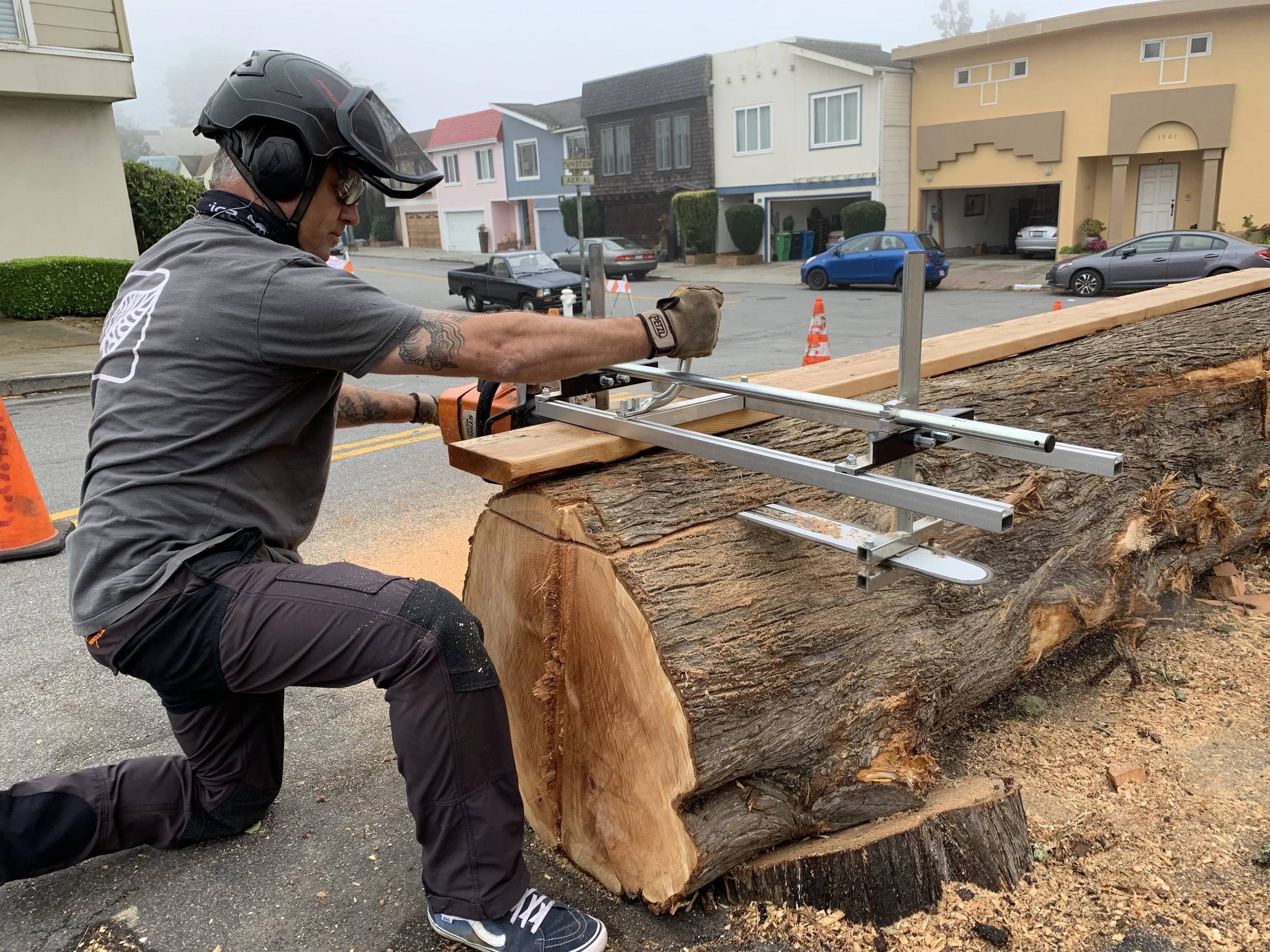 Man in helmet using a chainsaw to cut a large fallen tree on a residential street.