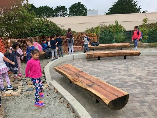 Children at a park sitting on benches and walking, with trees and a building in the background.