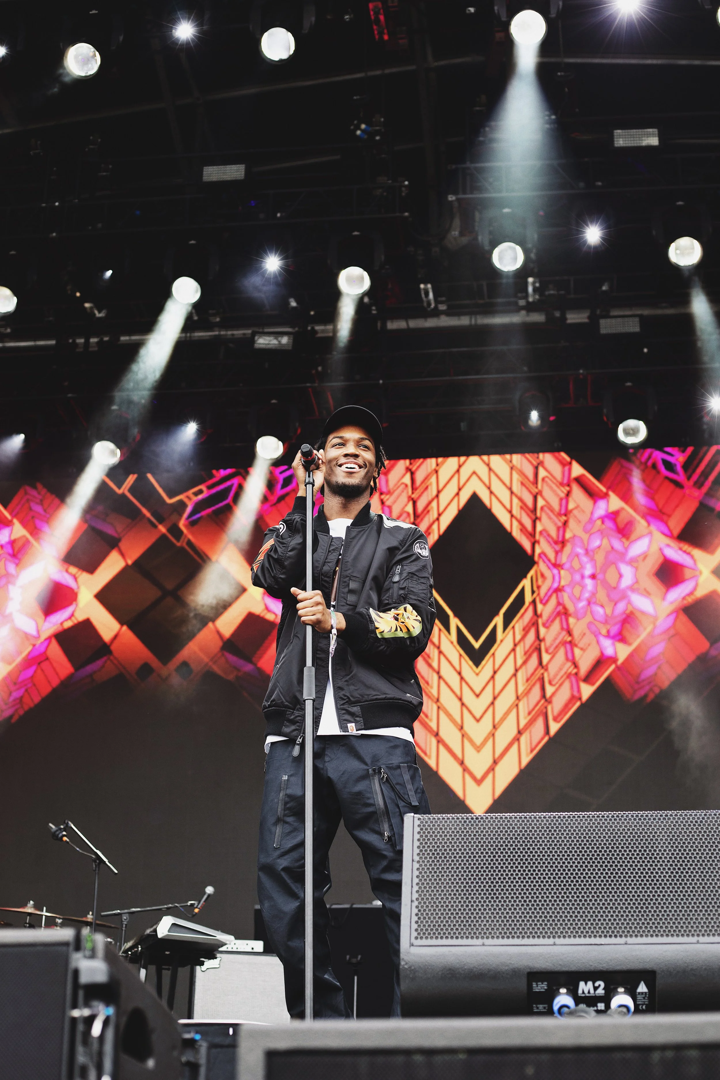 A male singer performs on stage during a concert, smiling while holding a microphone. The background features a colorful geometric pattern and stage lighting.