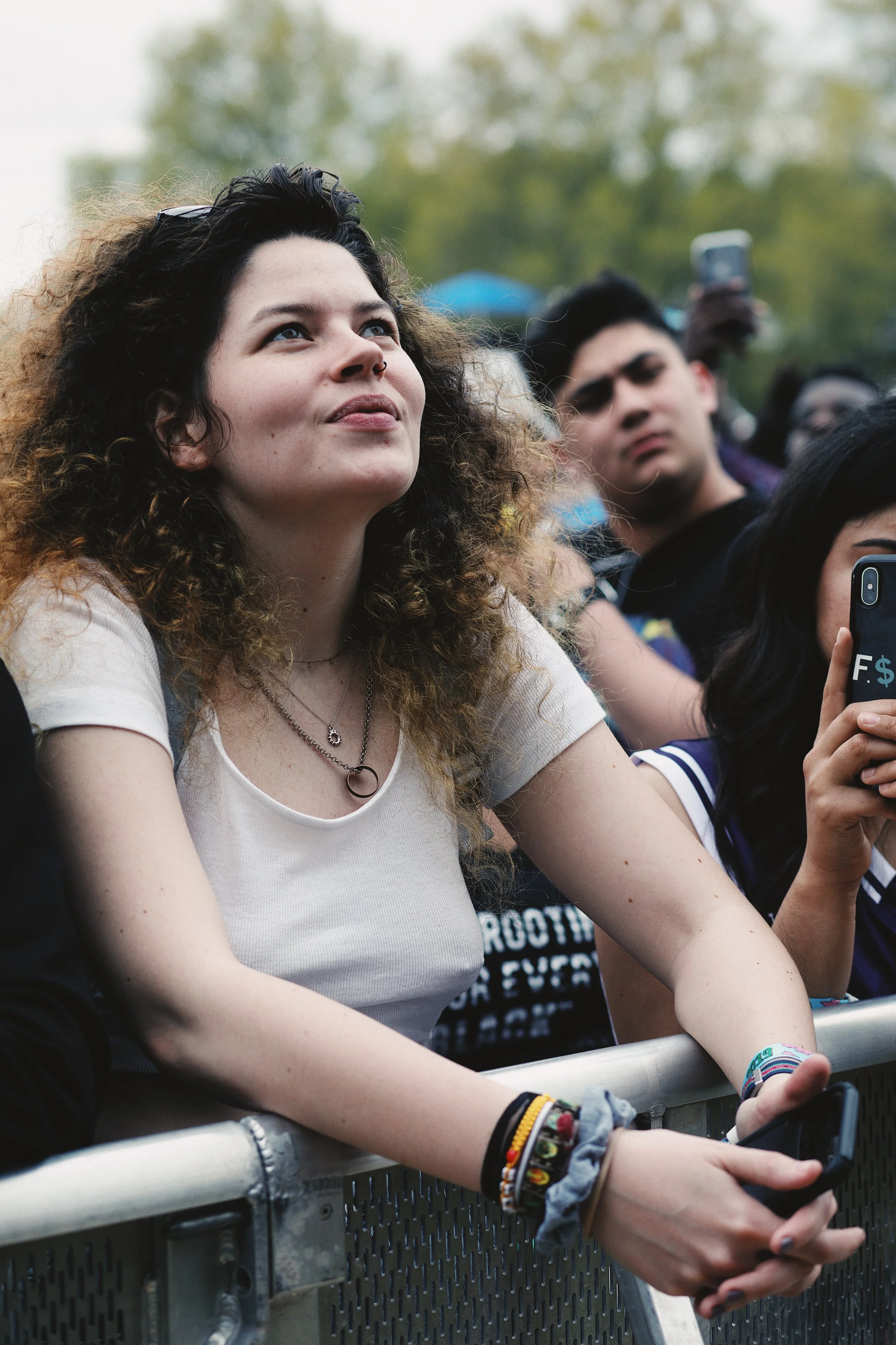 A young woman with curly brown hair and a septum piercing at a concert, leaning on a metal barrier with her hands clasped, holding a phone. Behind her, other concert attendees are visible, some holding phones, with trees in the background.