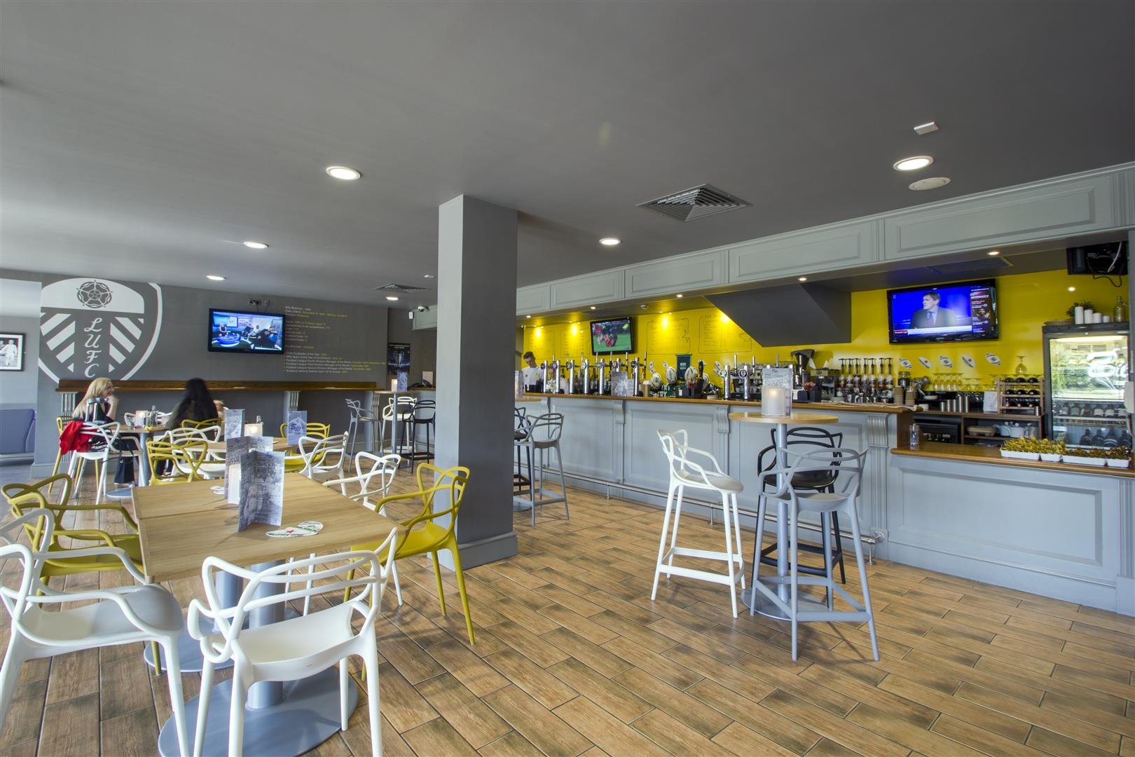 Billy’s Bar seating area and bar counter, with wood-effect floor, panelled grey bar front, yellow back-bar wall, TV screens and a Leeds United crest wall graphic