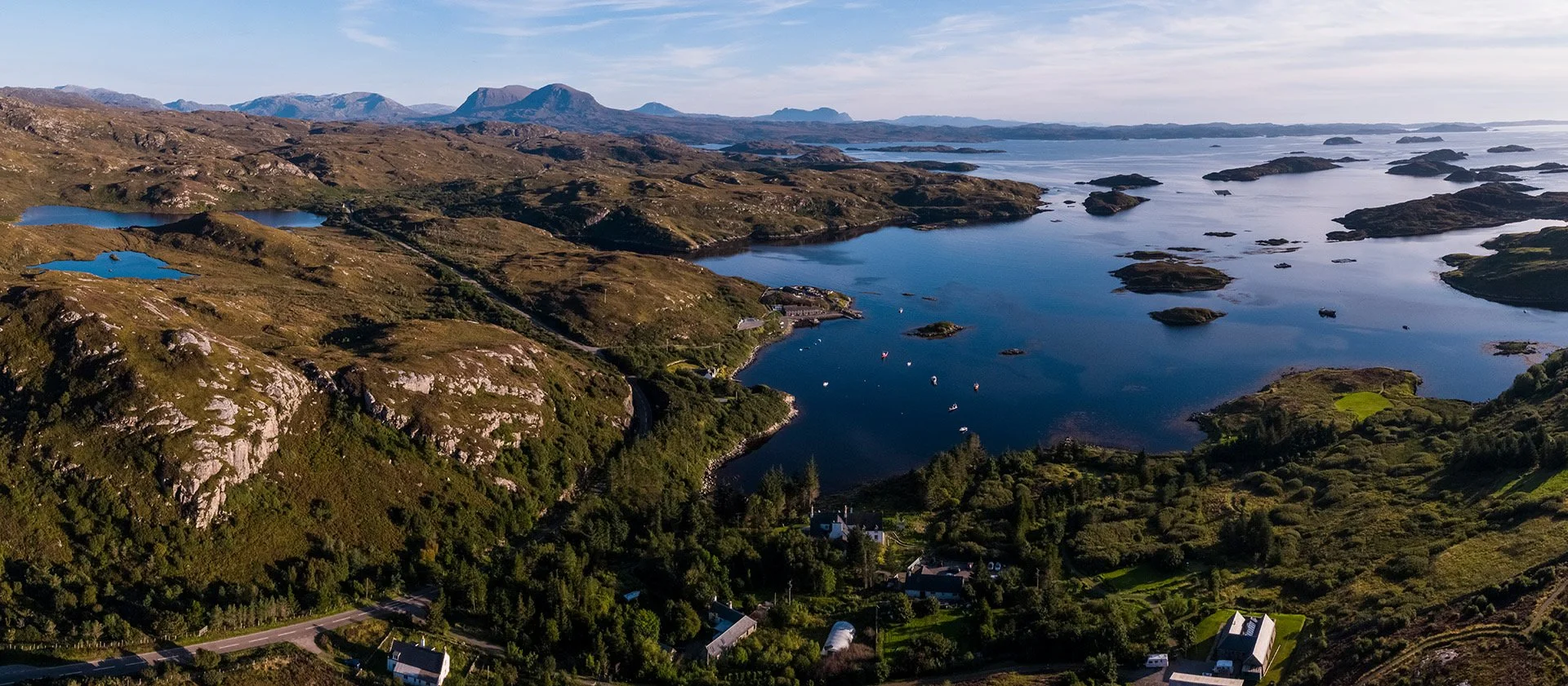 Eddrachill Hotel overlooking Badcall Bay with islands and skerries studded in the blue water. In the dsitance the hills of Assynt  and to the left the North Coast 500 cuts across the rocky landscape.