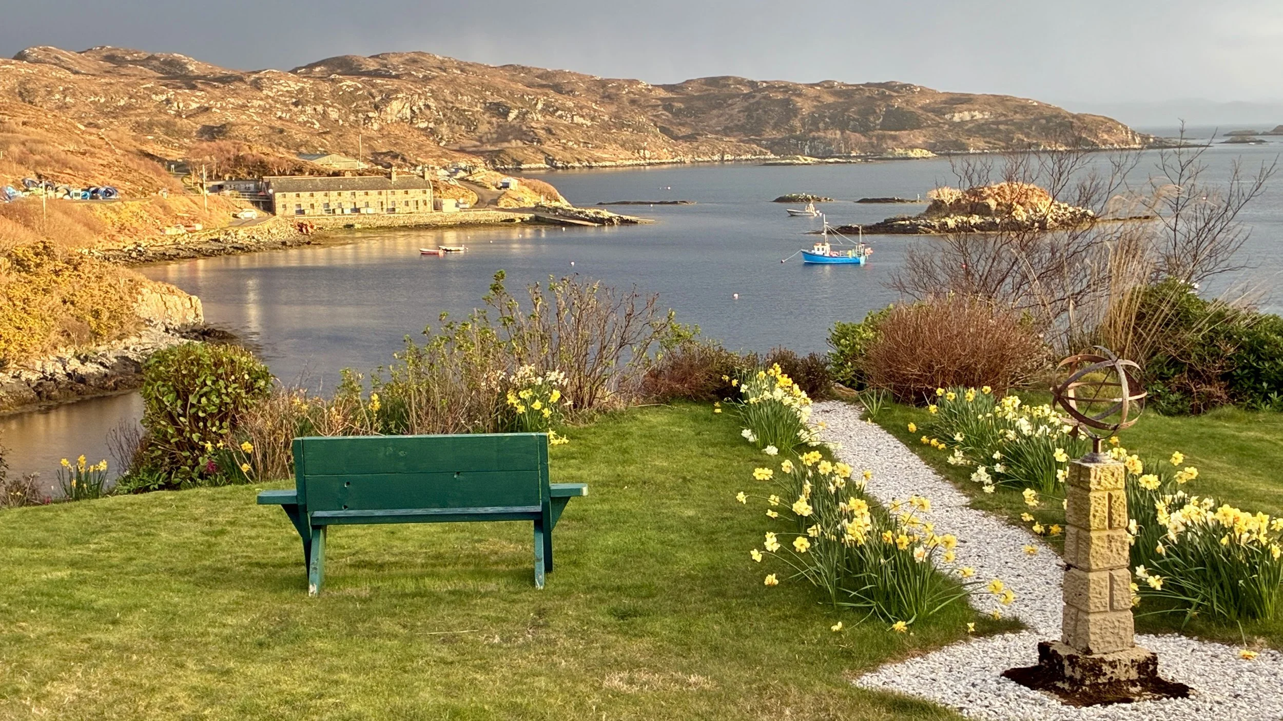 from the hotel lauwn in early evening, light blue fishing boat moored in middle distance,  green painted bench in foreground, white gravel path with sundial in centre. Daddodils in bloom. Hills still brown with fallen bracken, new growth