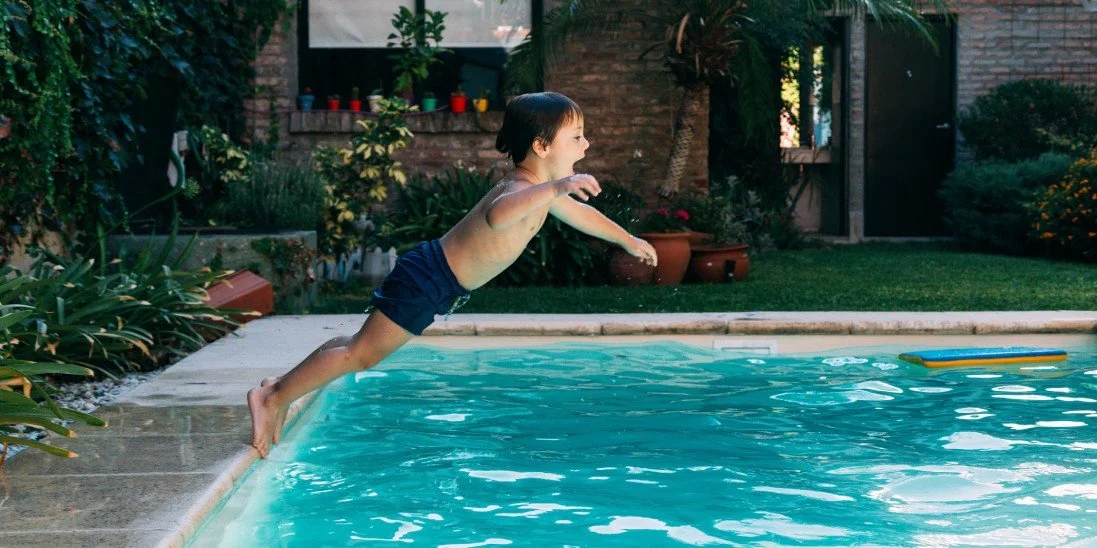 A little boy jumping into his family pool in the backyard. He's wearing swim shorts and a big smile as he jumps.