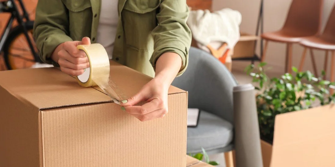 A person wearing a green jacket uses a masking tape to seal a carton, packing up potted plants and furniture.