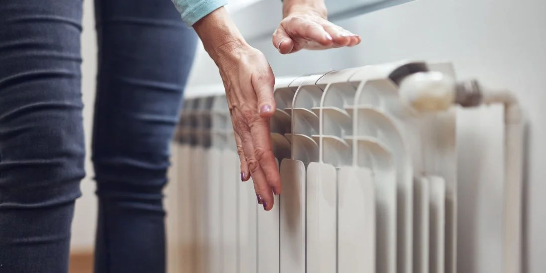 A homeowner stands next to their wall radiator as they place their hands near the vent tops to check the temperature.