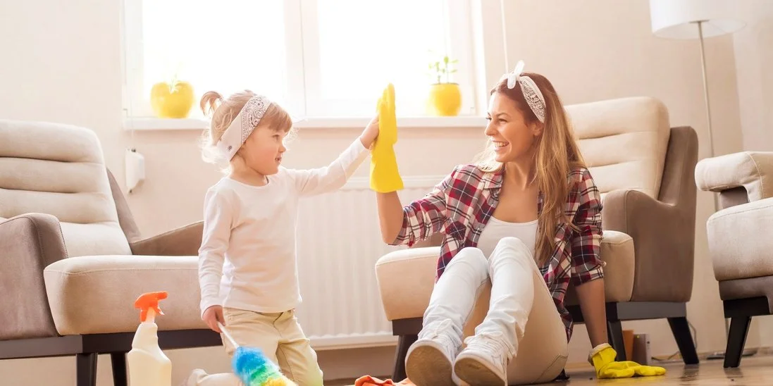 A child holding a feather duster high fives a woman wearing yellow cleaning gloves. They are wearing matching bandanas.