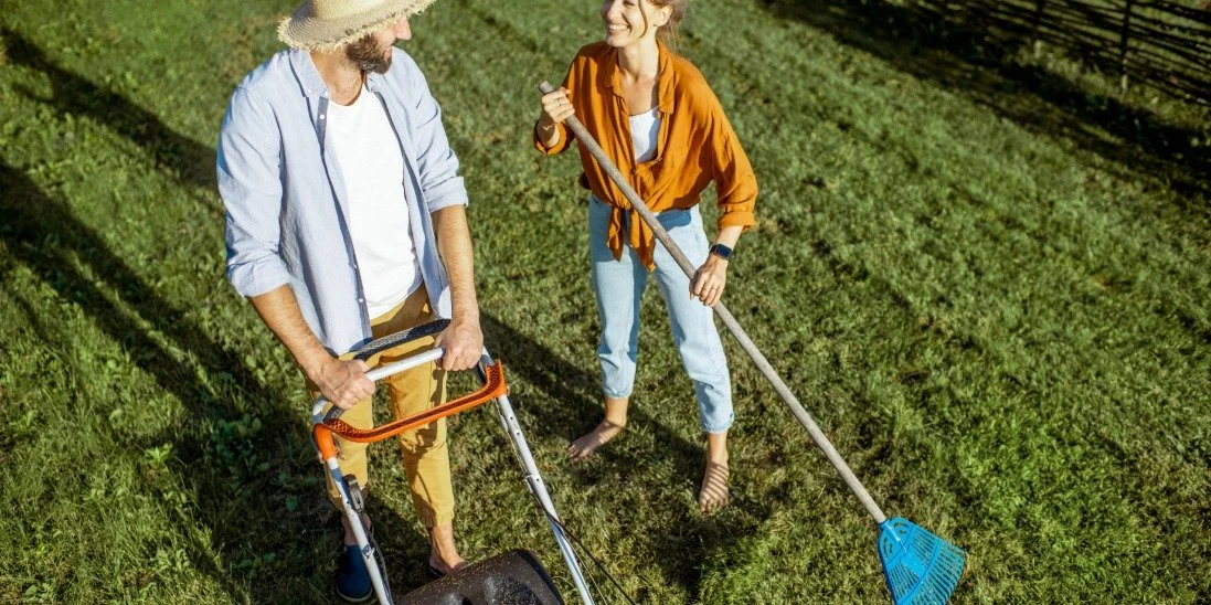 Man and woman work in a backyard, mowing the lawn and raking leaves together on a bright, tidy green lawn.