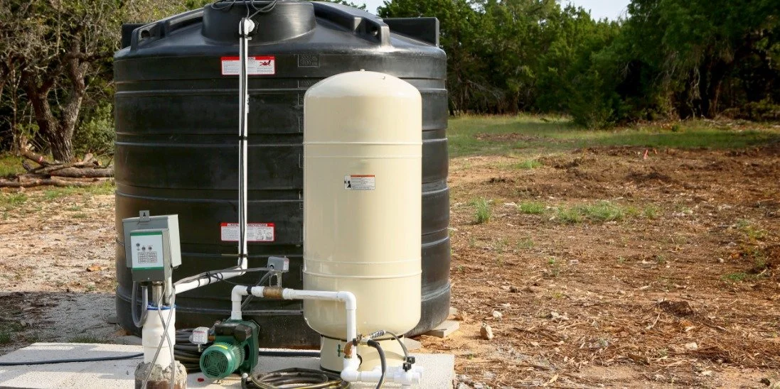 A black water storage tank stands beside a white pressure tank and pump on a concrete pad near a wooded yard.