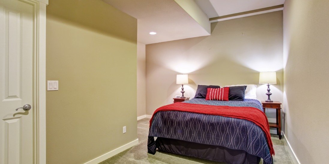 A small basement bedroom with a blue and red queen-sized bed in the middle. There are two nightstands with lamps turned on.