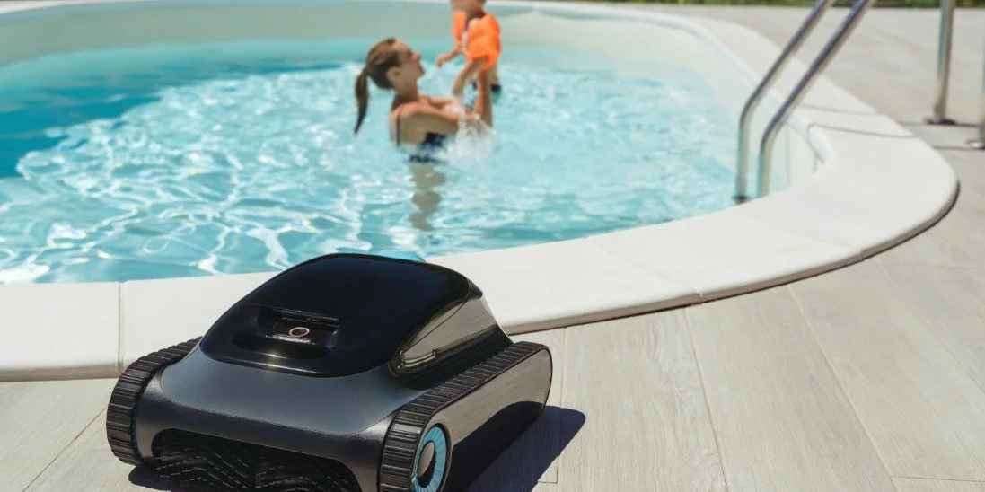 A robotic cleaner next to the edge of a swimming pool. A mother and her child are in the pool in the background.