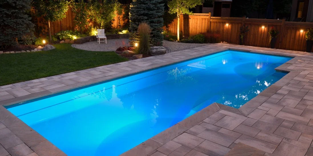 An in-ground pool surrounded by a concrete paver deck at nighttime. Lights inside the pool cause the water to glow.
