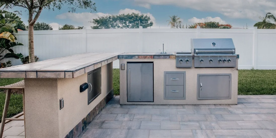 A modern outdoor kitchen with built-in grill, sink, storage cabinets, and bar beside a white privacy fence.