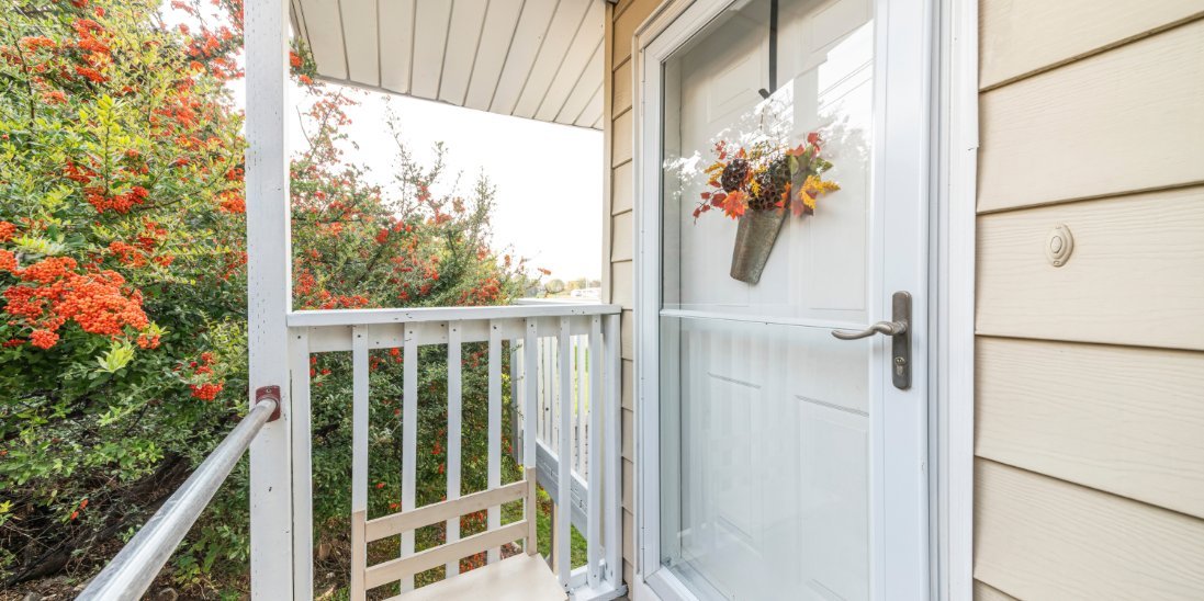 A front porch with a white storm door, railing, chair, and flowering shrubs beside a beige-sided home entrance.