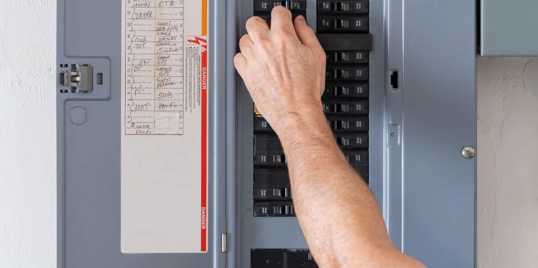 An individual adjusting a circuit breaker inside an open electrical panel with labeled switches and breaker directory.
