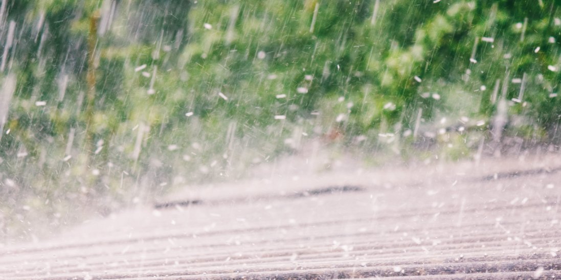 A roof is being pelted with rain and hail. The roof is made of slate and there is greenery in the background.