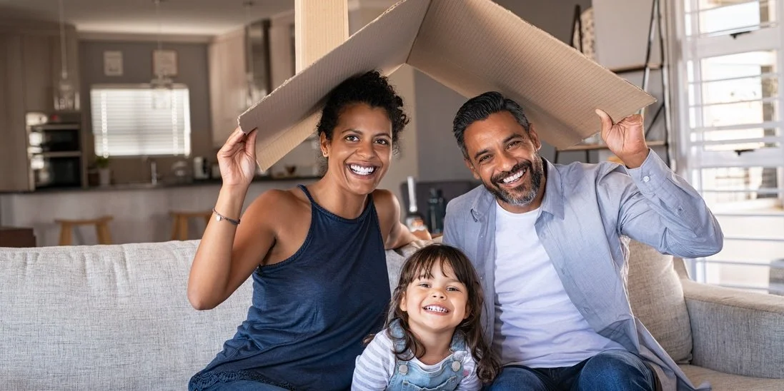 A man and a woman sits on the sofa and holds a cardboard mimicking a roof. In between them is a younger child.
