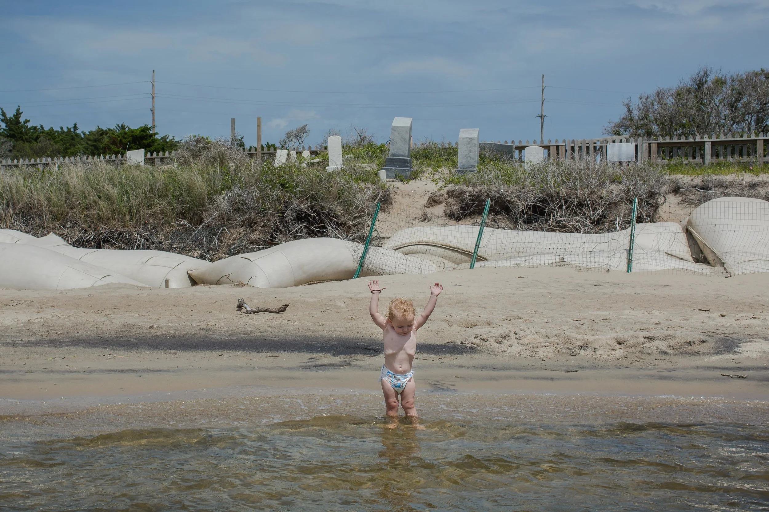 Kaine, 2, plays in the sound near sandbags installed to break waves during storms and slow erosion in the cemetery. May 2018.