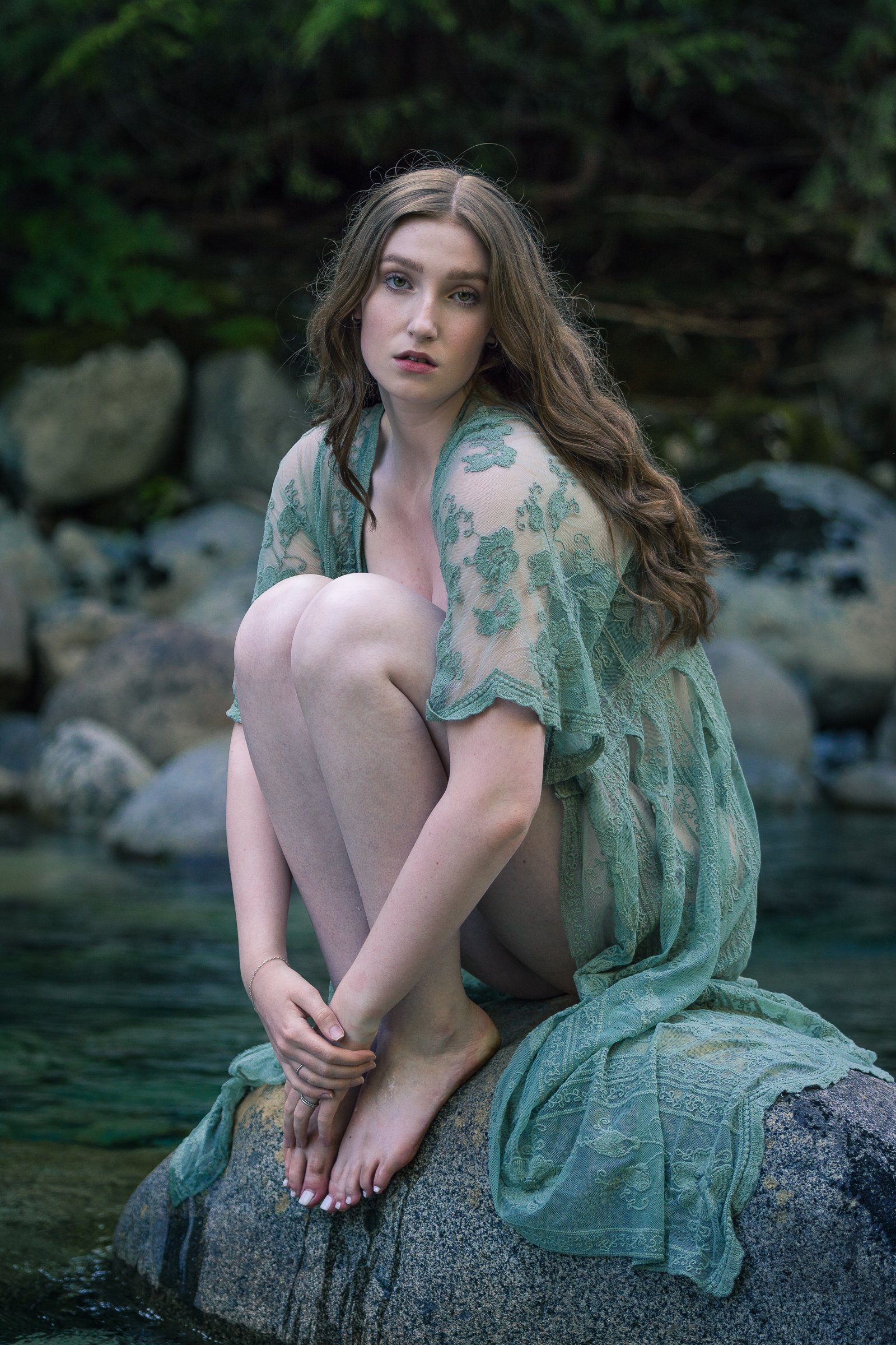 A young woman with long brown hair and light eyes, wearing a sheer green embroidered dress, sitting on a rock in a river with lush green foliage in the background.