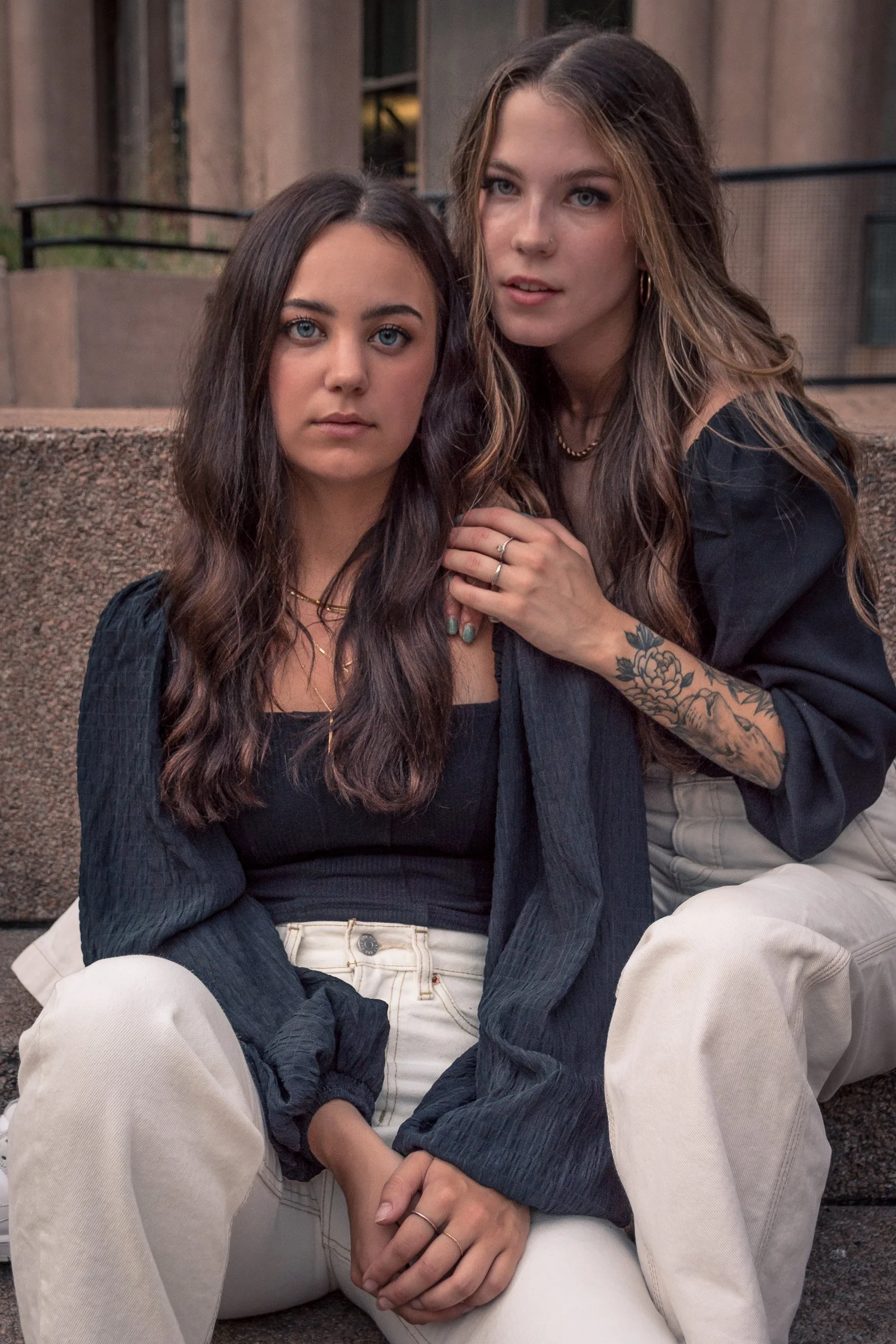 Two young women sitting close together outdoors with a concrete building behind them. One has long dark hair and blue eyes, the other has long light brown hair, blue eyes, and tattoos on her arm. Both are wearing casual clothing and looking at the ca