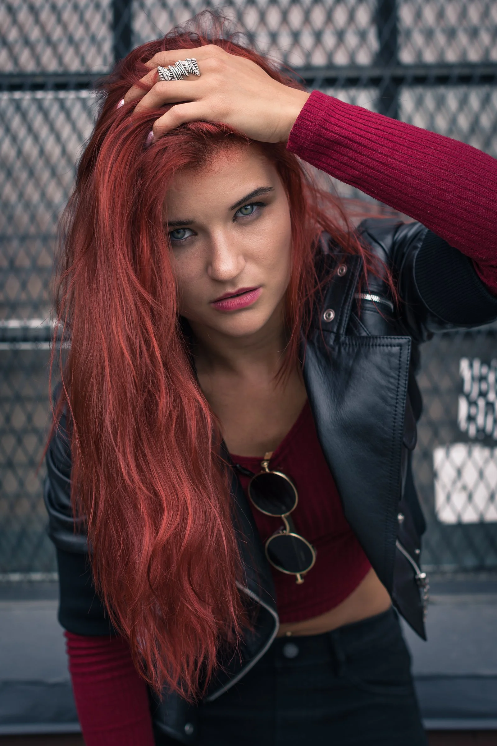 A young woman with long red hair, blue eyes, and fair skin, wearing a black leather jacket and a red top, posing in front of a chain-link fence, with one hand on her head and looking at the camera.