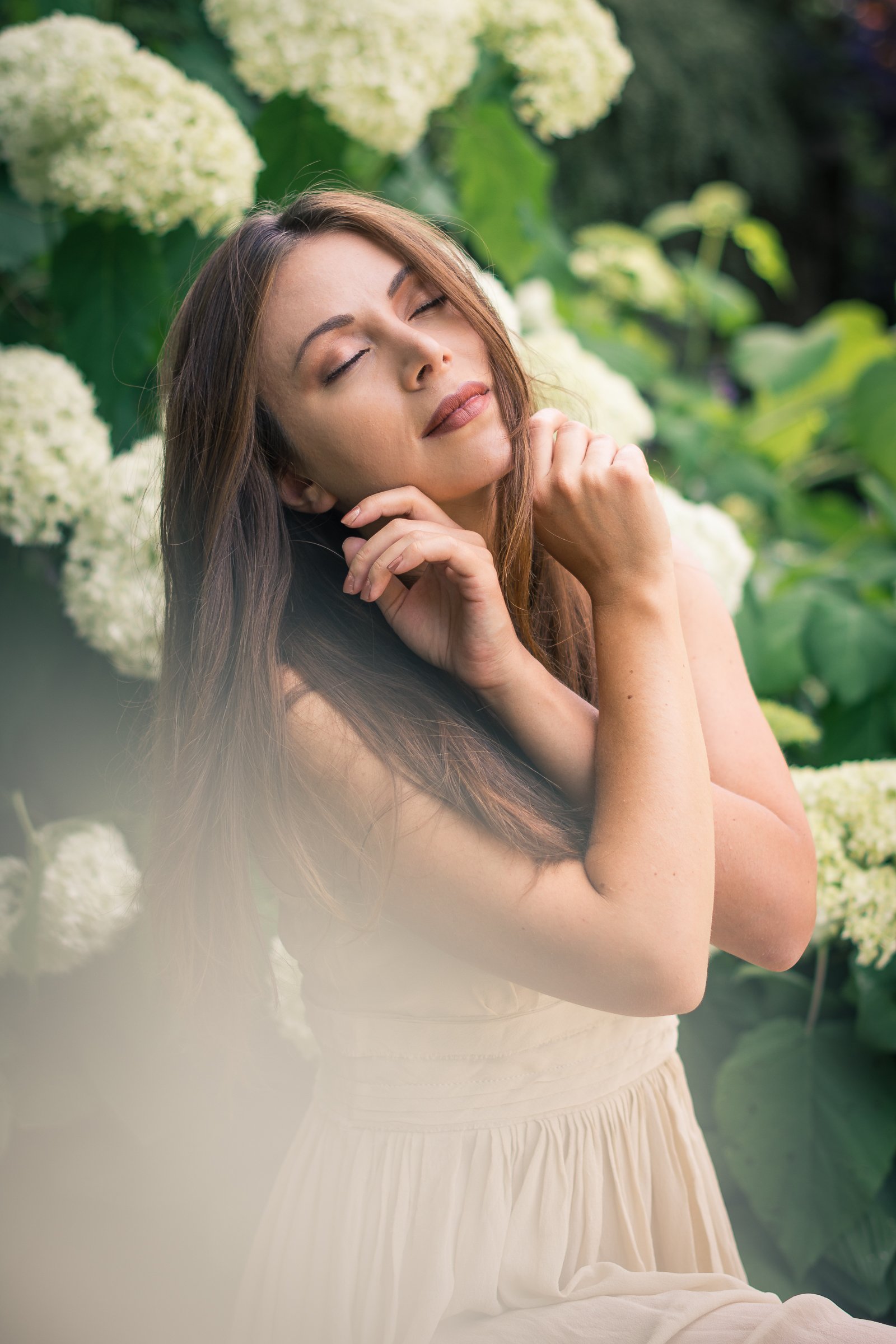 A woman with long brown hair and closed eyes, smiling gently, standing outdoors surrounded by green foliage and white hydrangea flowers, with her hands near her face.