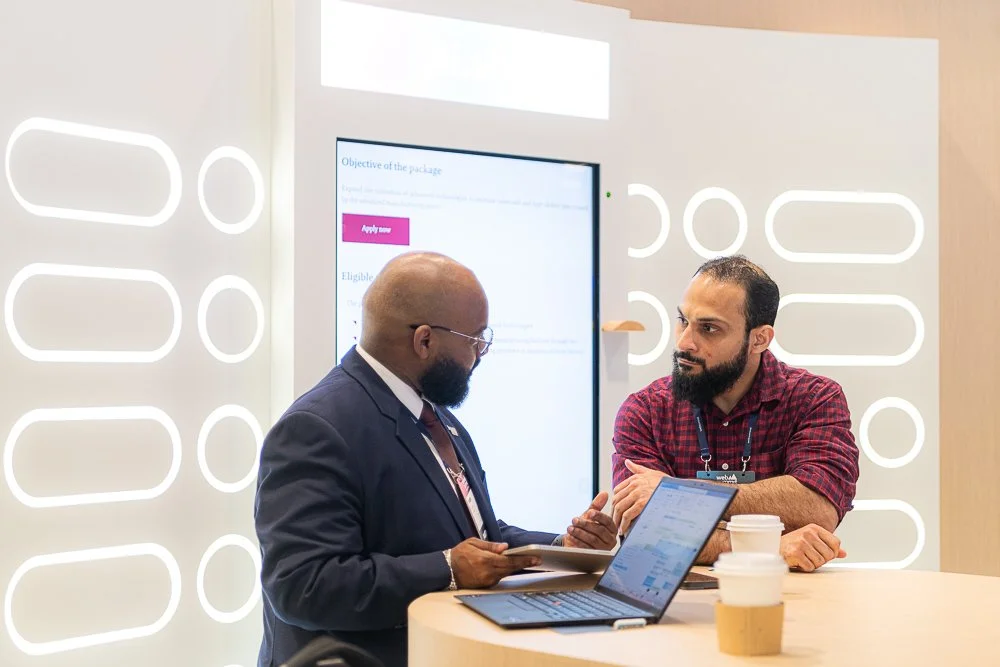 Two men are having a discussion at a table with laptops and coffee cups in a modern, brightly lit office or conference space.