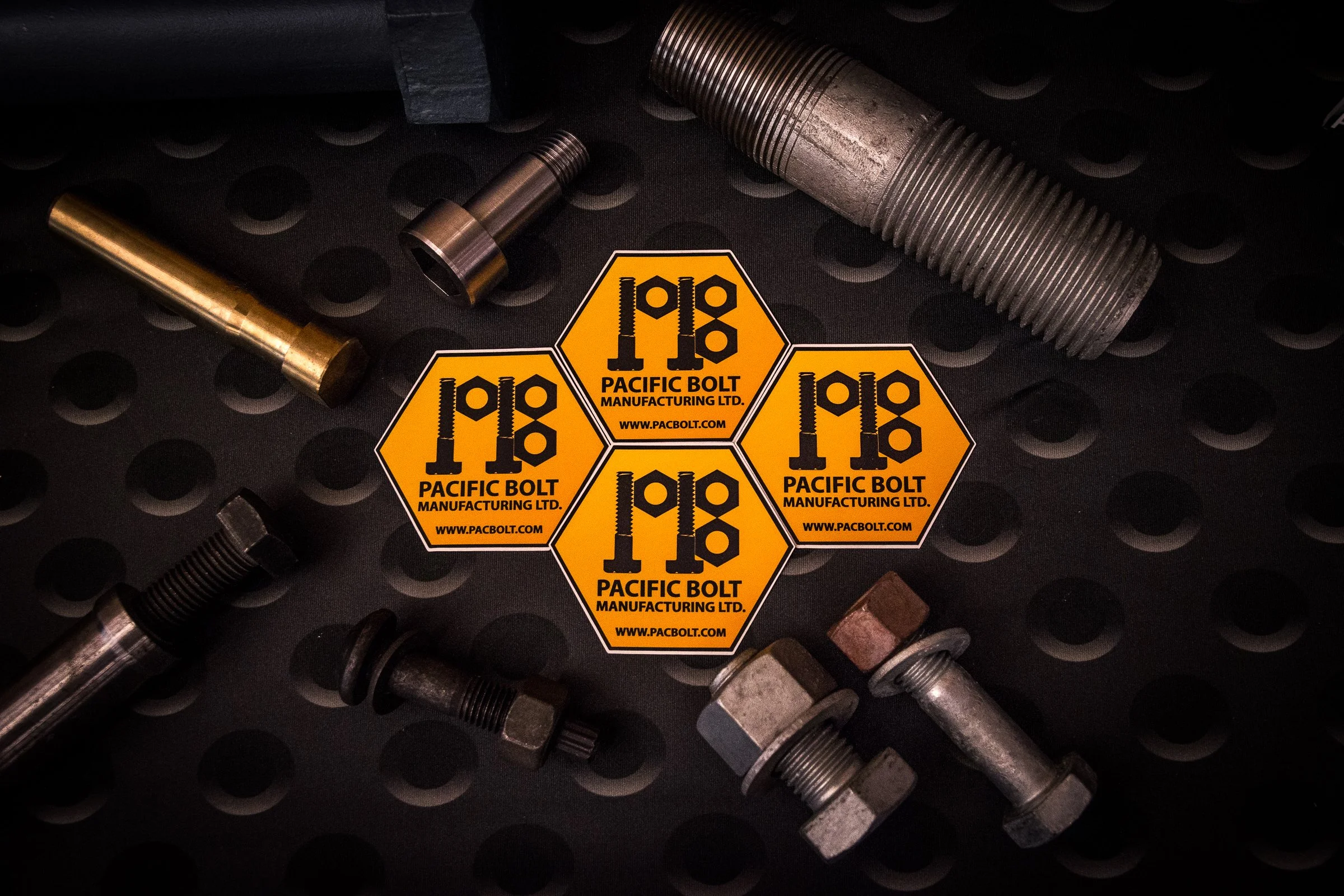 Various metal bolts and fasteners, including a large threaded bolt, a smaller threaded bolt, and some spacers or bushings, are arranged on a black textured surface. There are also four yellow stickers from Pacific Bolt Manufacturing Ltd.
