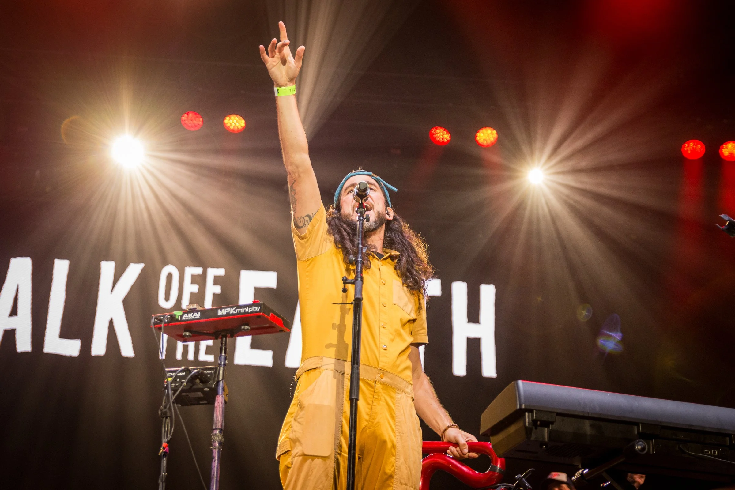 A male musician with long hair, a beard, and tattoos, wearing a blue bandana and yellow shirt, performs on stage with his arm raised. There are bright stage lights and musical equipment around him.
