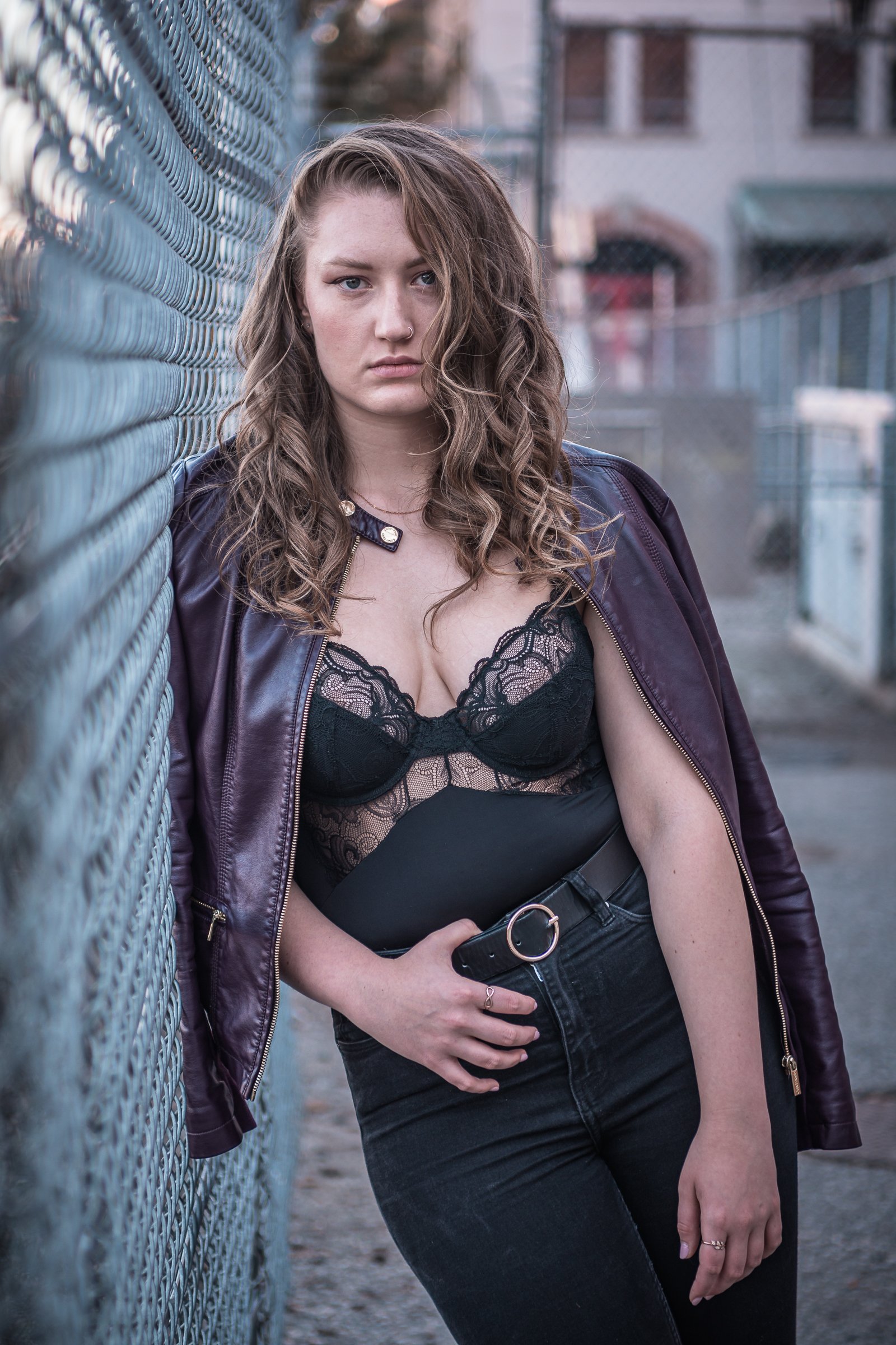 A young woman with long, curly brown hair leaning against a chain-link fence in an urban setting, wearing a black lace top and a purple jacket hanging off her shoulders, holding a black clutch and wearing a belt with a gold round buckle.