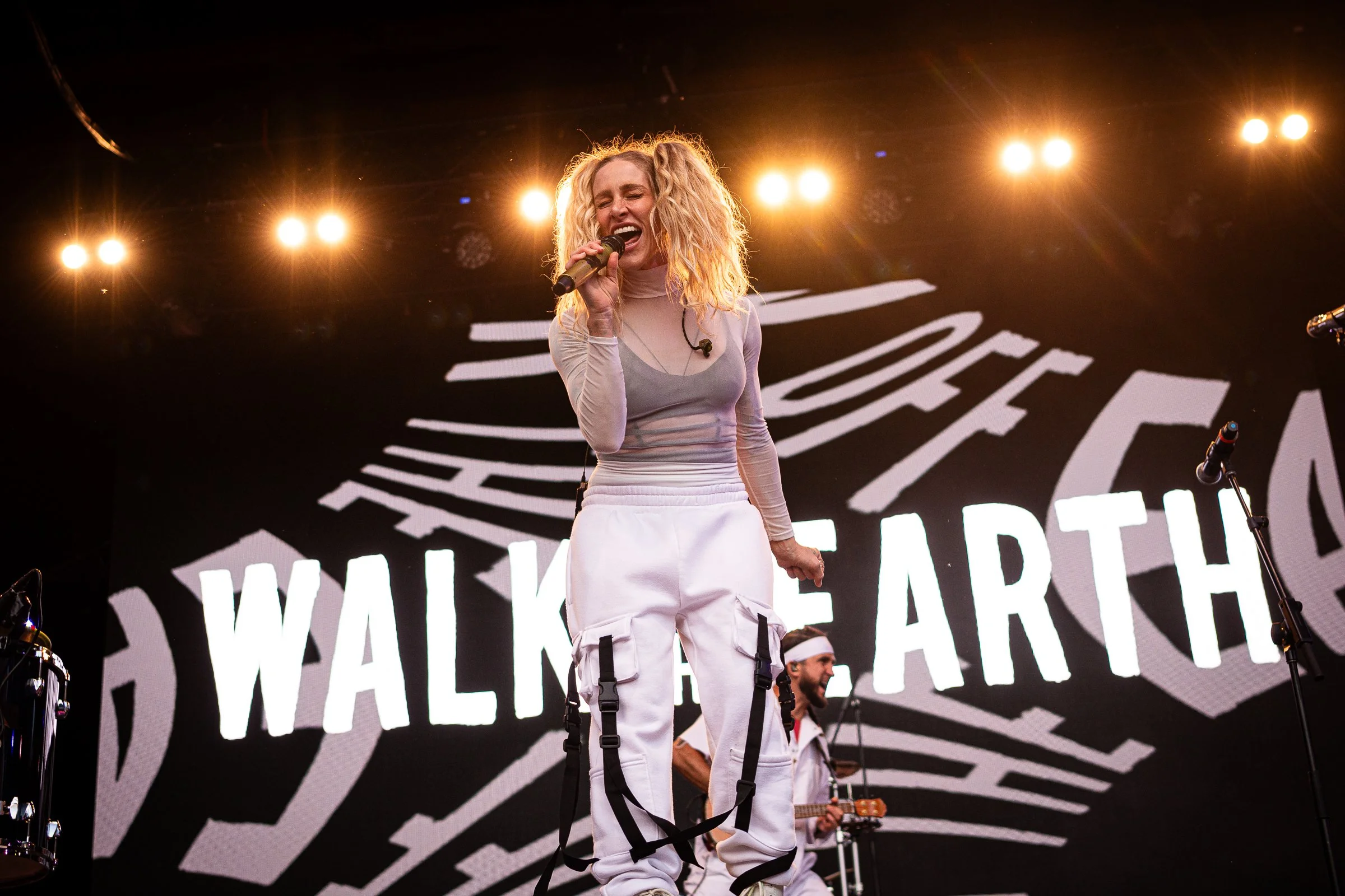 Female singer performing on stage with bright lights behind her, wearing a light-colored long-sleeve top and white cargo pants, holding a microphone and singing passionately.
