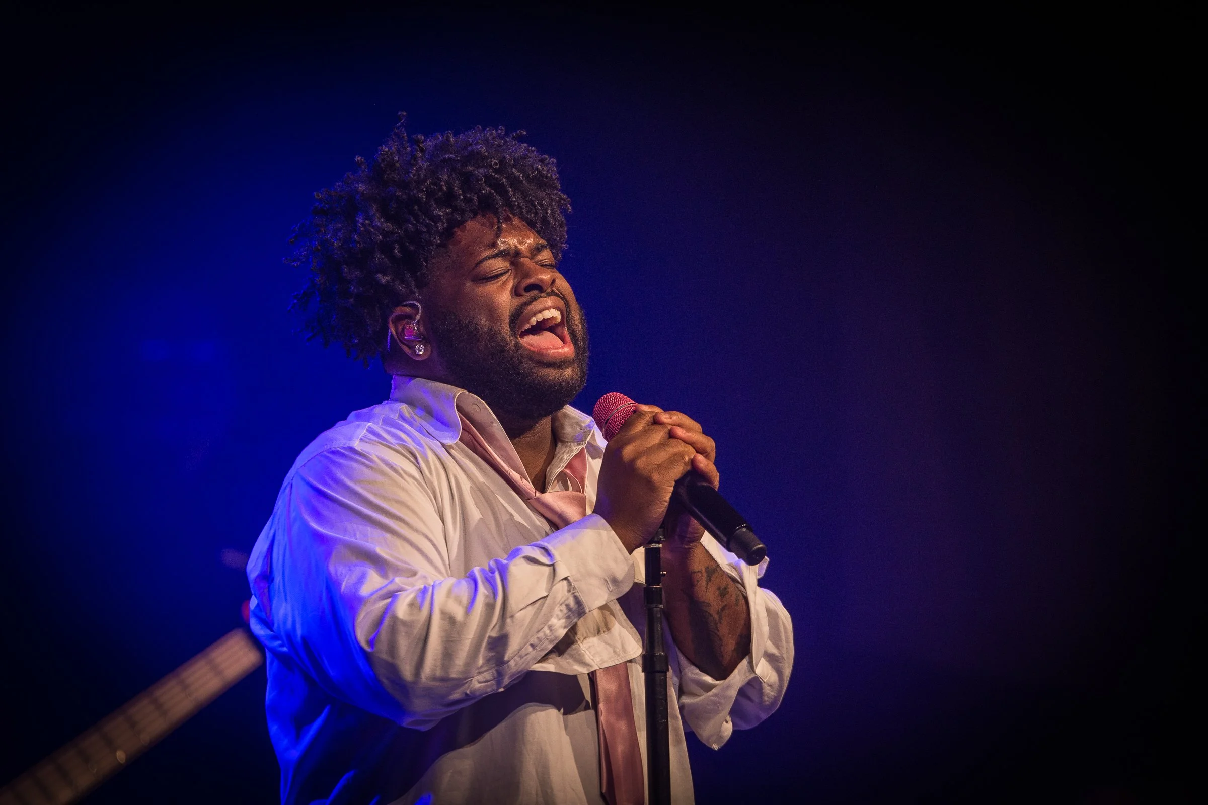 A male singer passionately performing on stage, holding a microphone with both hands, eyes closed, wearing a white shirt with rolled-up sleeves, illuminated by blue stage lighting.