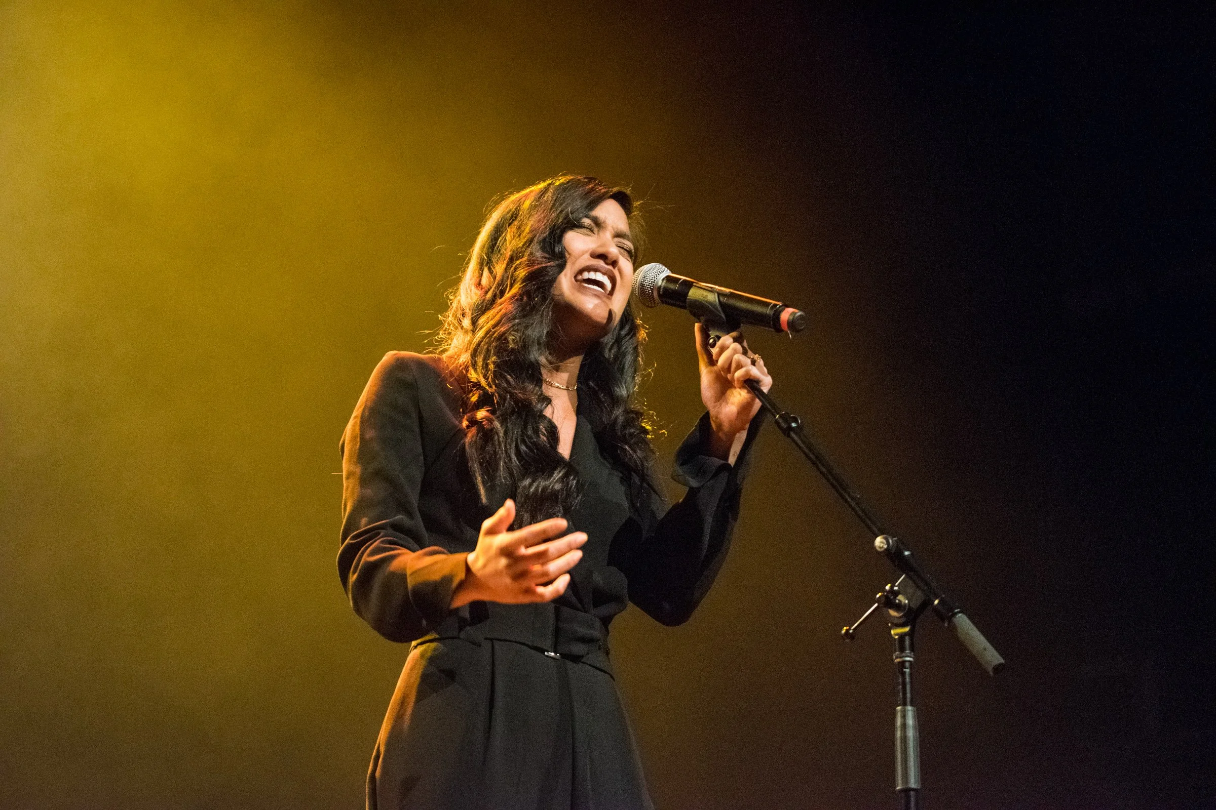 A woman with dark, wavy hair singing passionately into a microphone on stage, dressed in a black outfit, with yellow stage lighting behind her.