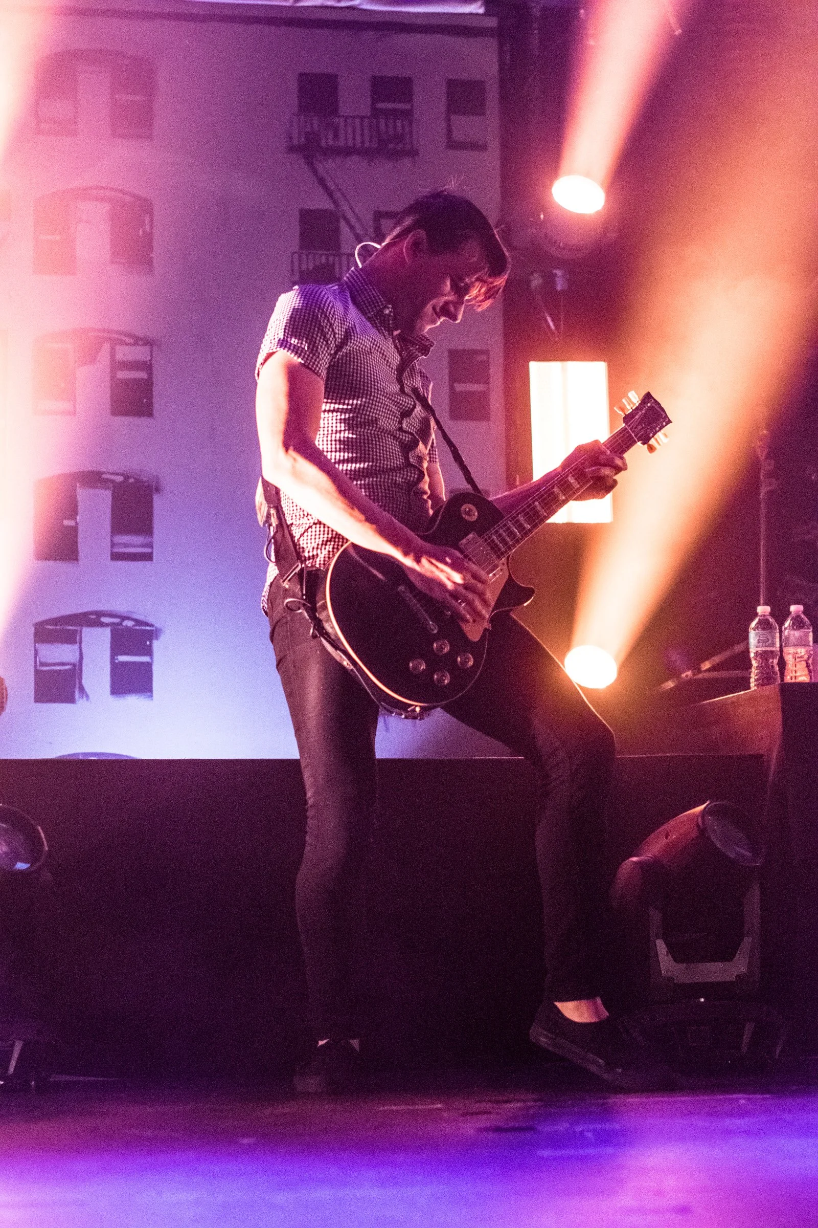 A person playing an electric guitar on stage under colorful stage lights, with water bottles on a nearby table and a backdrop featuring building windows.