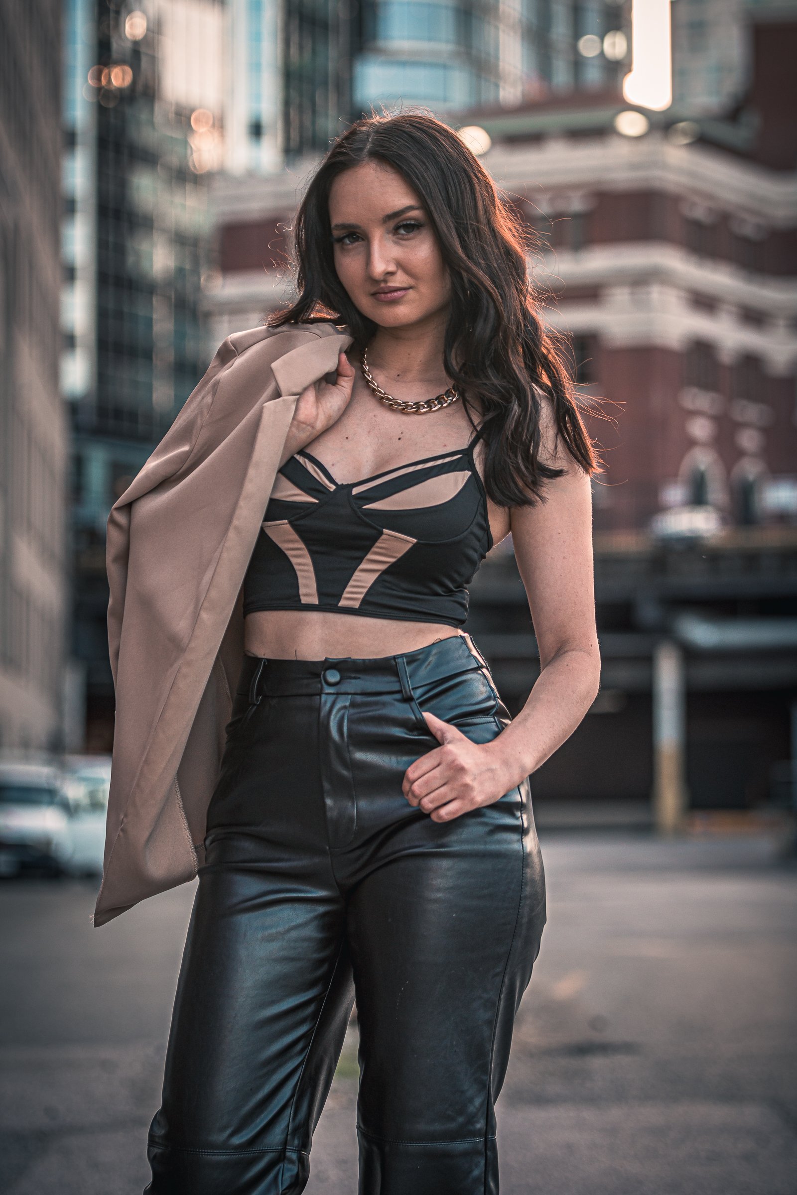 A young woman with dark wavy hair, wearing a black crop top, black leather pants, a beige jacket draped over her shoulder, and a gold chain necklace, stands in an urban setting with buildings and city lights in the background.