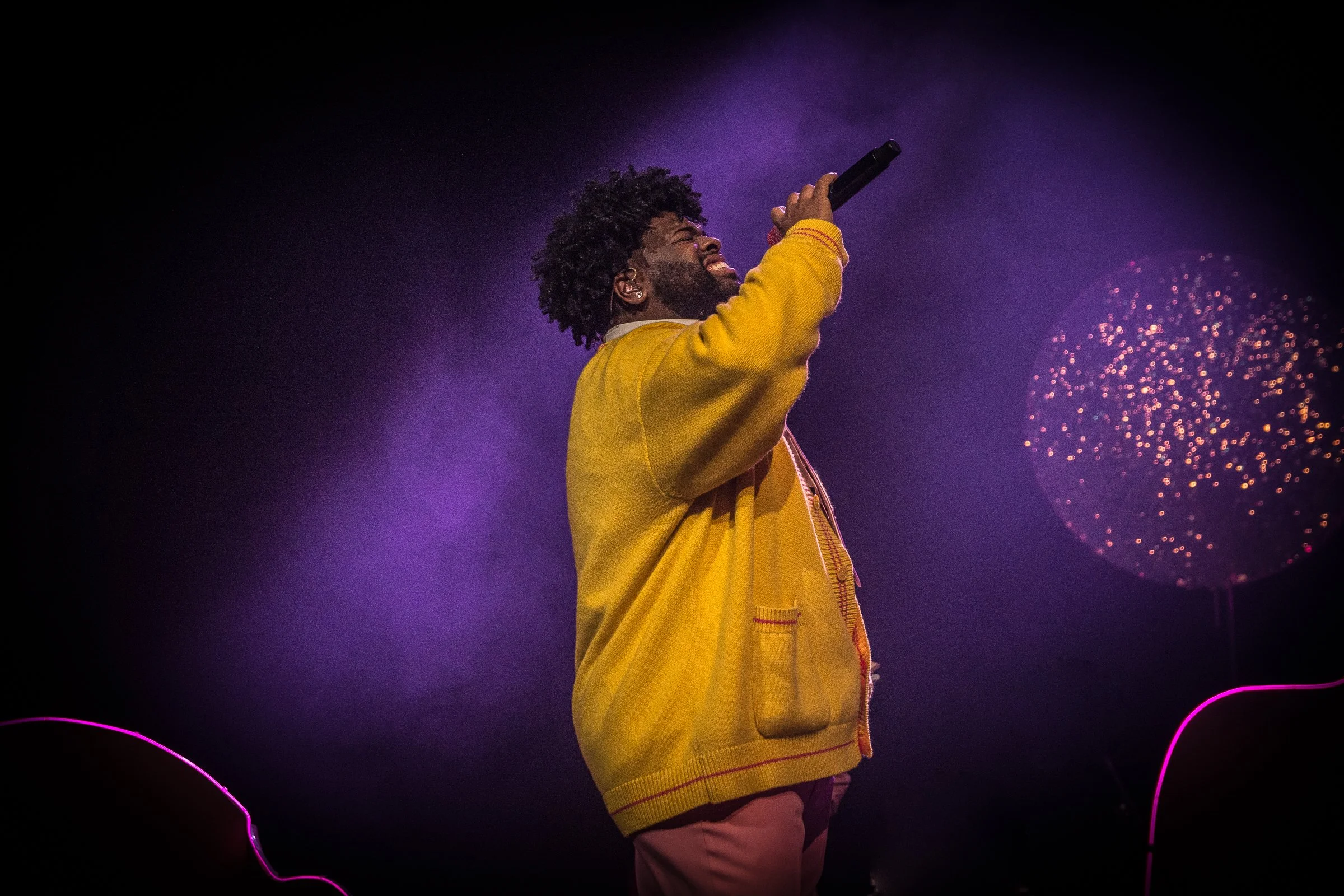 A man with curly hair and a beard wearing a yellow jacket singing into a microphone on stage with purple lighting and fireworks in the background.