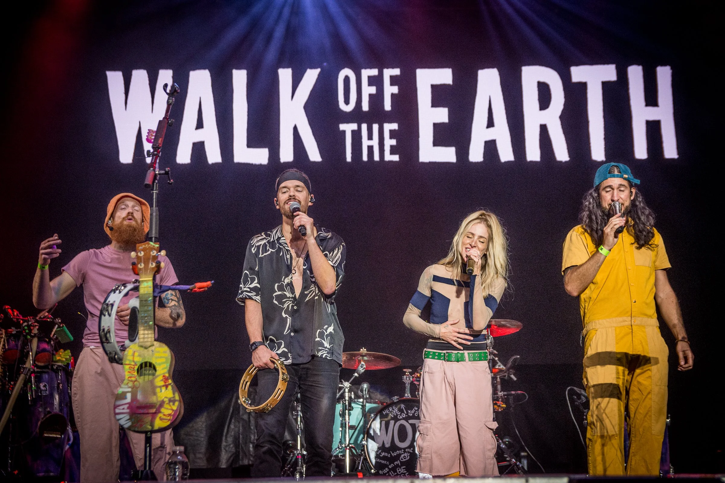 A group of five musicians on stage performing with a large screen behind them displaying the phrase 'WALK OFF THE EARTH' in bold white letters.