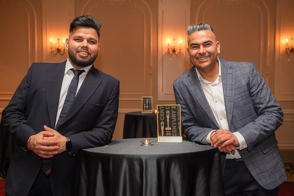 Two men in suits standing by a round table at a formal event, smiling. One has dark hair and beard, the other has gray hair. The background features chandeliers and elegant wall decor.