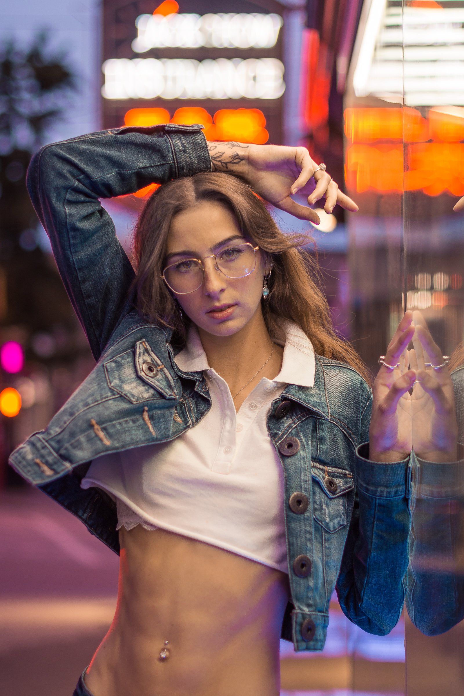 A young woman with glasses and long hair is posing in front of a neon-lit background, wearing a denim jacket over a white crop top, and has a belly button piercing.