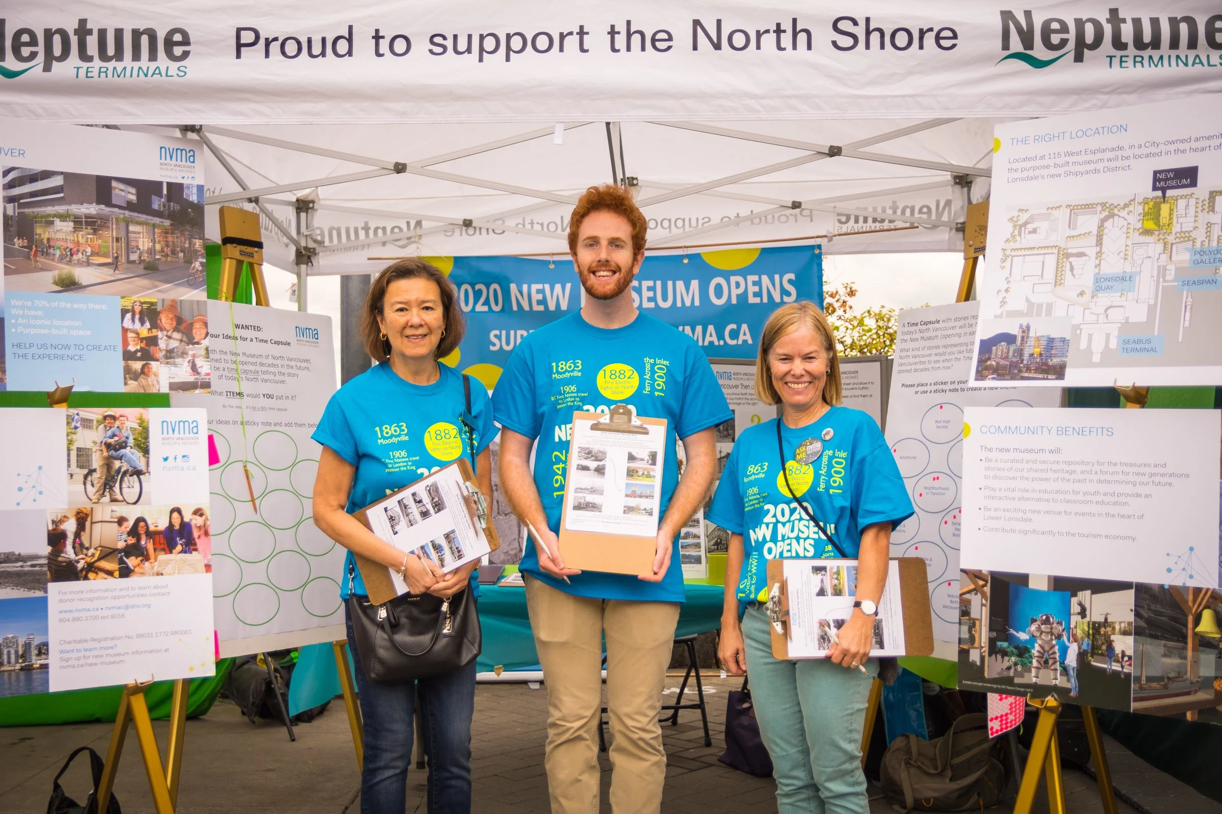 Three people standing at an information booth about a new museum opening, holding brochures and smiling. The booth has various informational posters and a banner that reads '2020 New Museum Opens'.