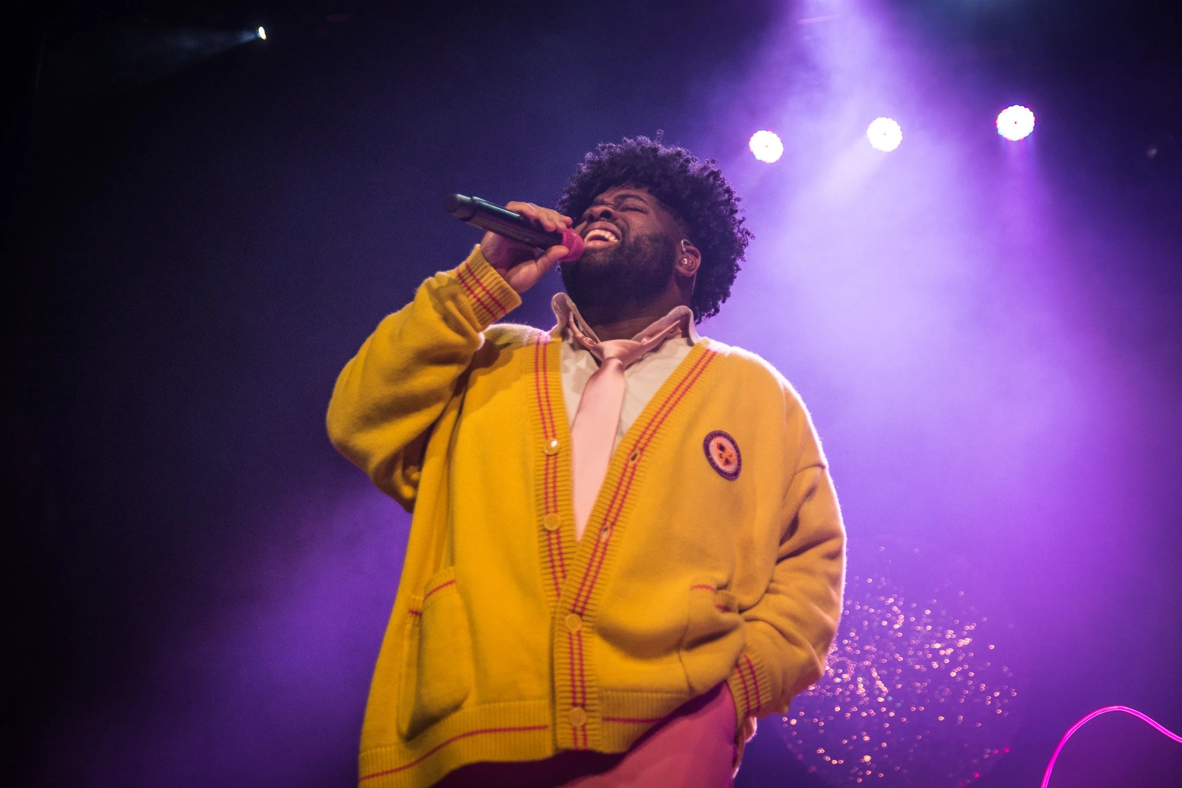 A man with curly hair and a beard singing into a microphone on stage, wearing a yellow cardigan with a pink shirt and pink tie, under purple stage lights.