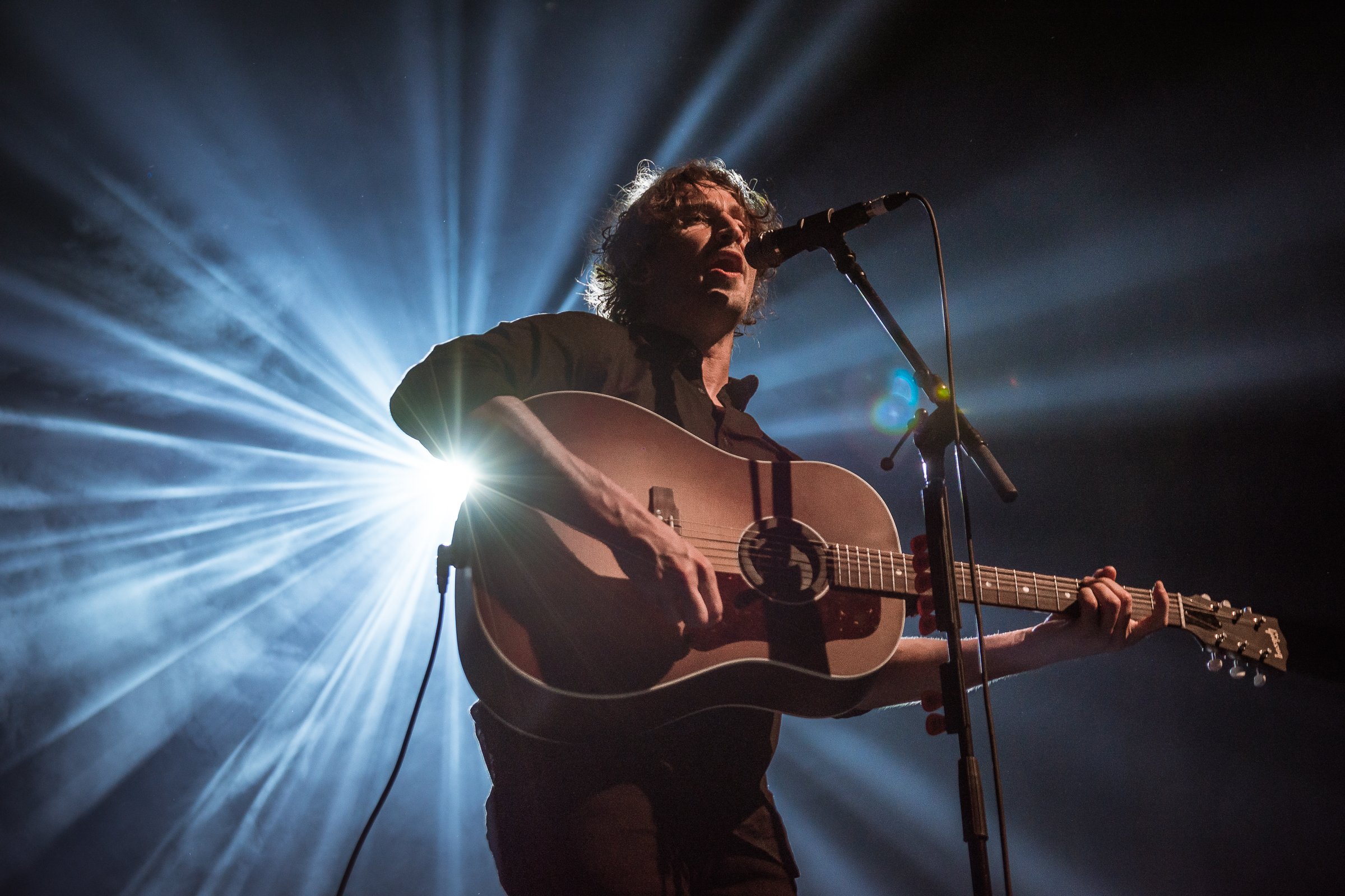 A man with curly hair playing an acoustic guitar and singing into a microphone on stage, illuminated by stage lights creating a starburst effect in the background.