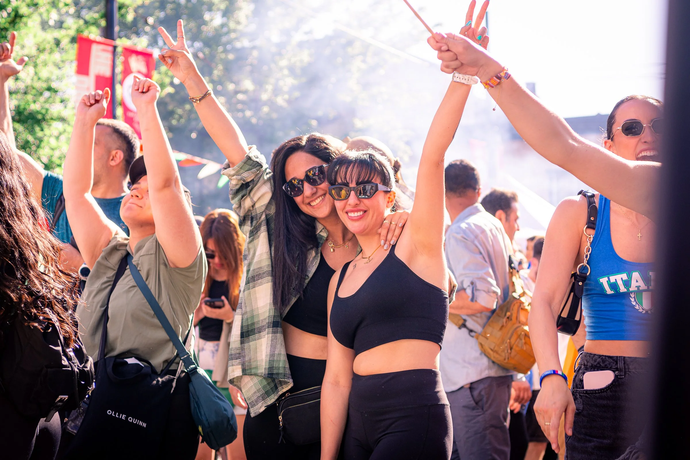 People celebrating outdoors on a sunny day, showing enthusiasm and happiness, some making victory signs with their hands, in a lively crowd.
