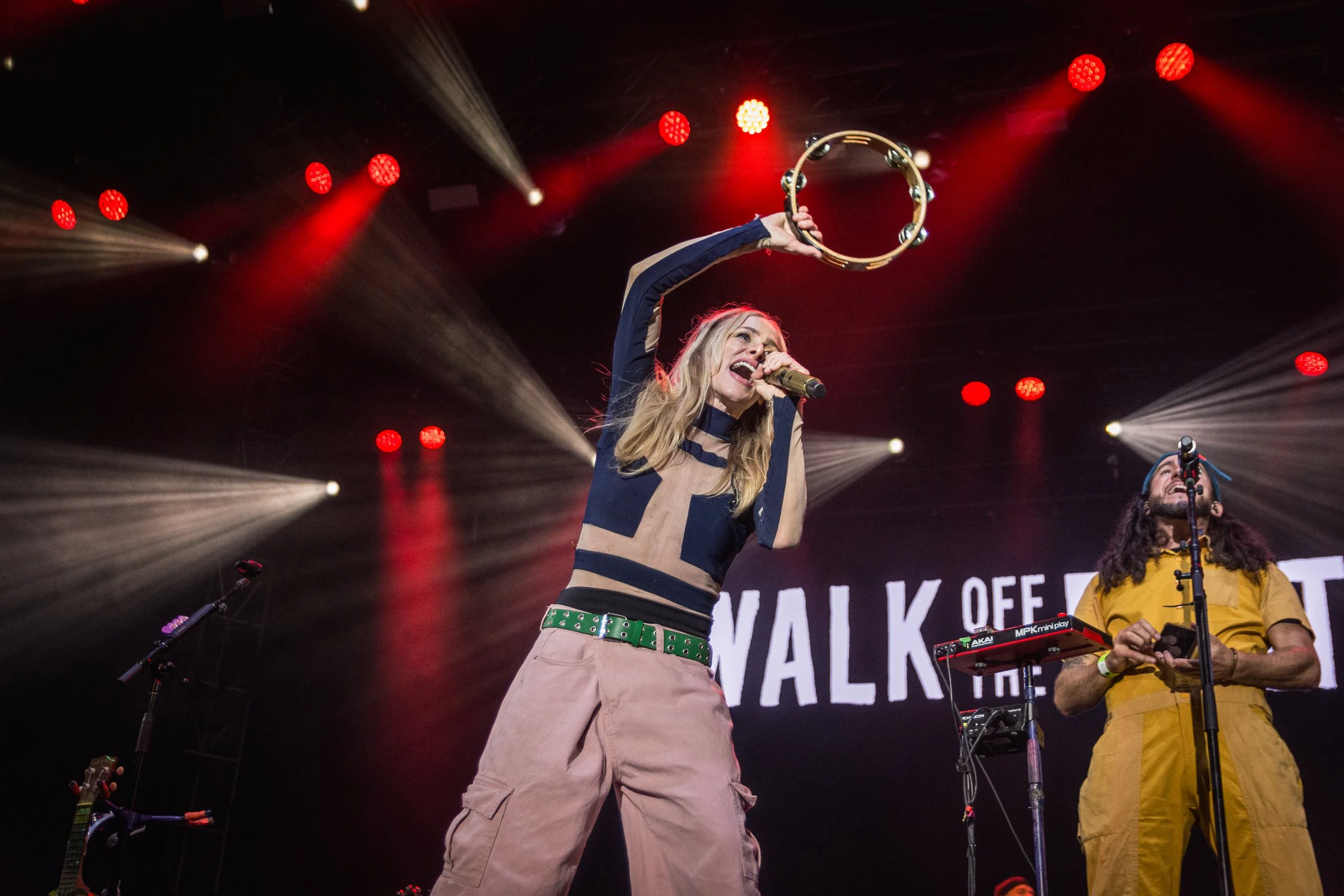 A woman with long blonde hair singing into a microphone while holding a tambourine above her head at a concert. She is wearing a blue and beige top and beige cargo pants with a green belt. A man with glasses and a yellow shirt is seen in the backgrou