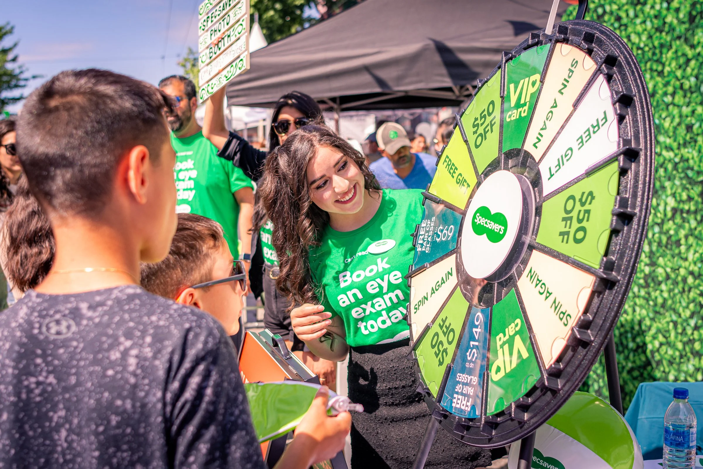 A group of people gathered around a prize wheel at an outdoor event. The wheel has various sections with text such as "Free Gift," "$50 Off," "Spin Again," and "VIP Card." A woman with long dark hair and sunglasses is smiling as she spins or points a